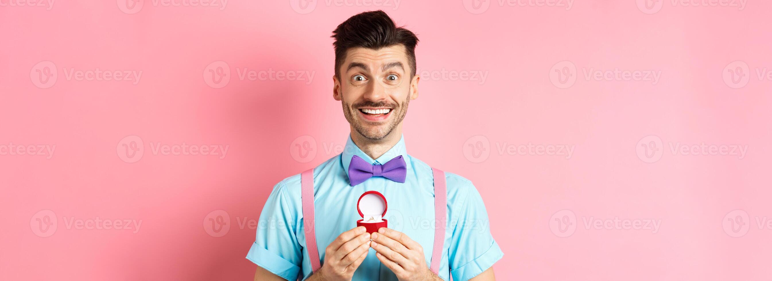Valentines day. Romantic man in bow-tie showing engagement ring and smiling, asking to marry him, making proposal to lover on pink background photo