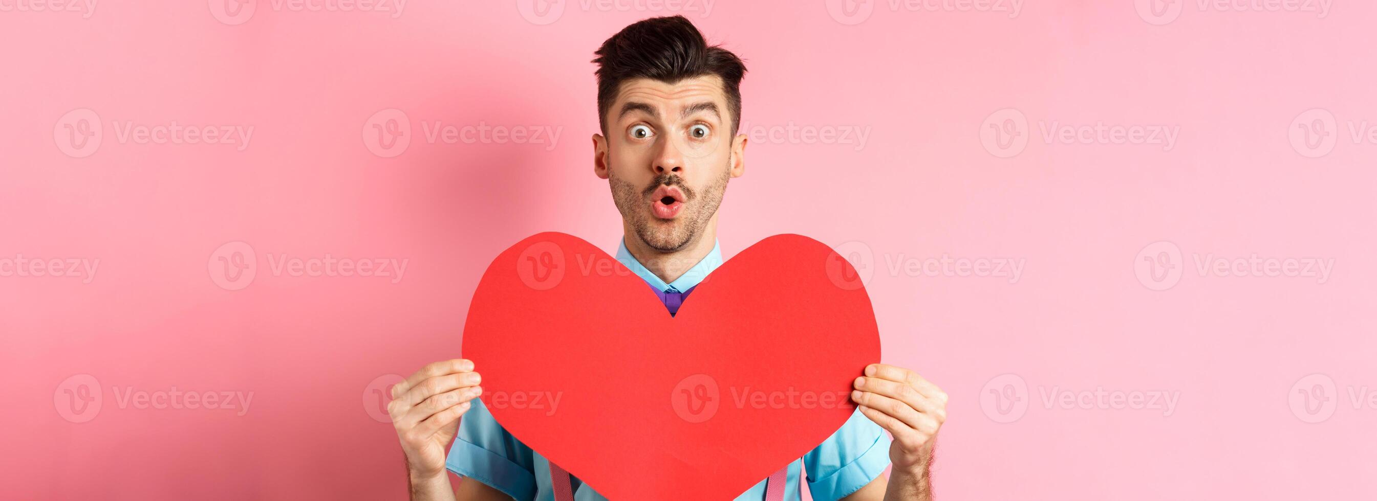 Valentines day concept. Amused young man with bow-tie and moustache, showing heart cutout and looking for love, standing on pink romantic background photo