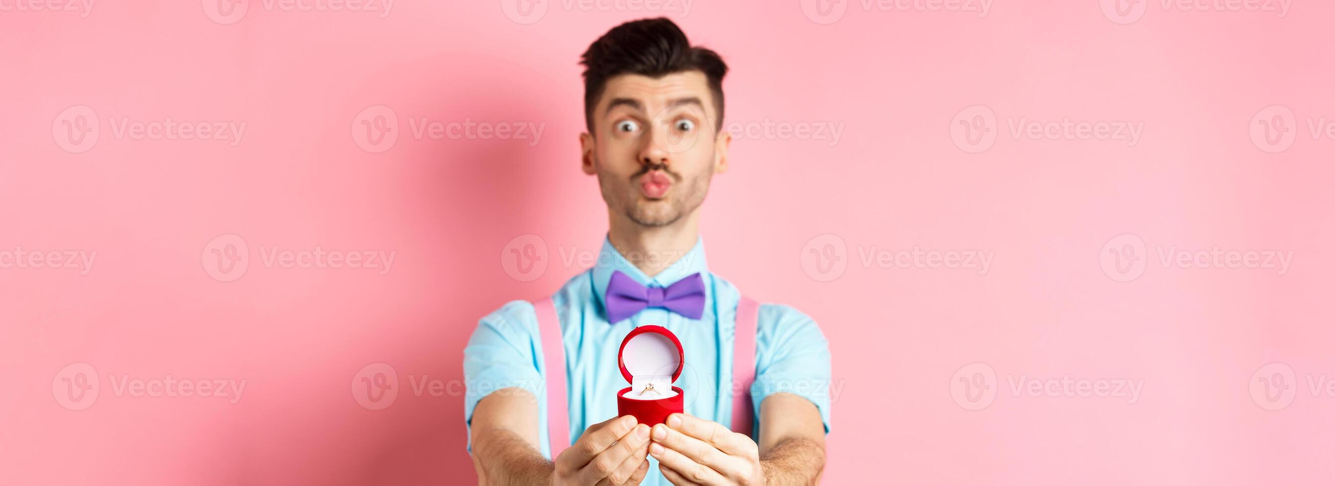 Valentines day. Funny young man pucker lips for kiss and showing engagement ring, making proposal, say marry me to lover, standing over pink background photo