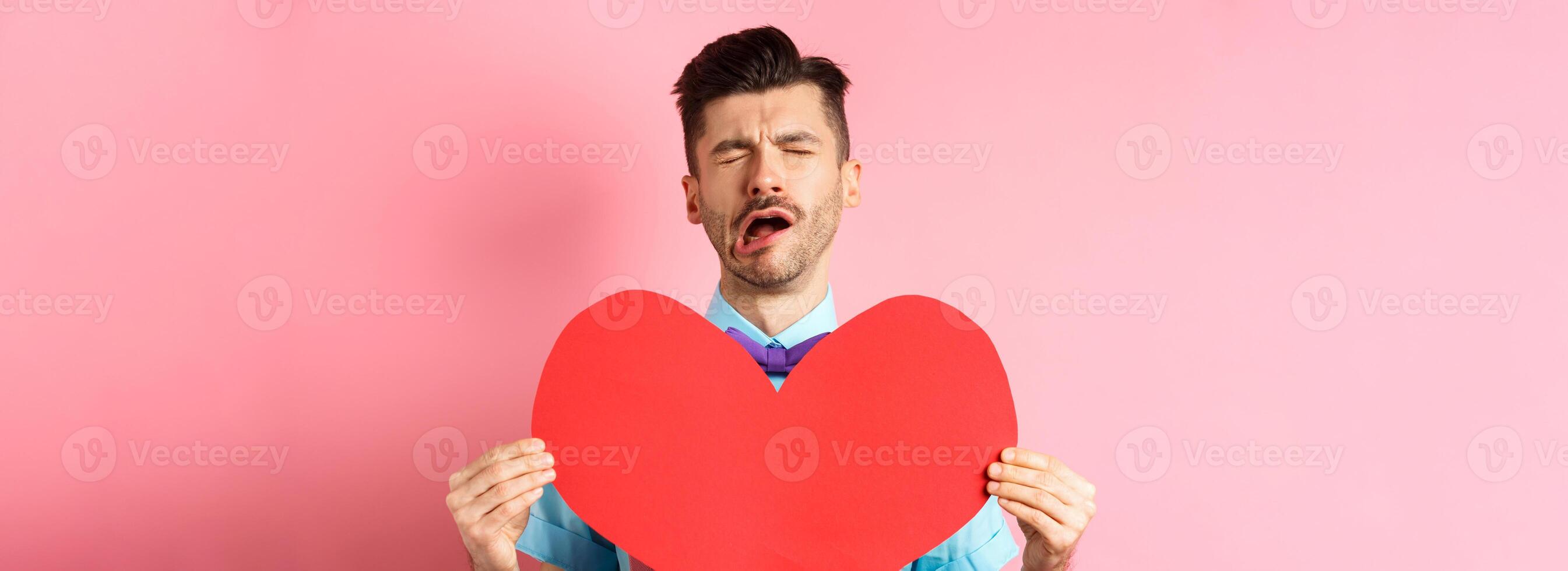 Valentines day concept. Sad and lonely man feeling heartbroken, being rejected, showing big red heart cutout and crying from break-up, standing on pink background photo