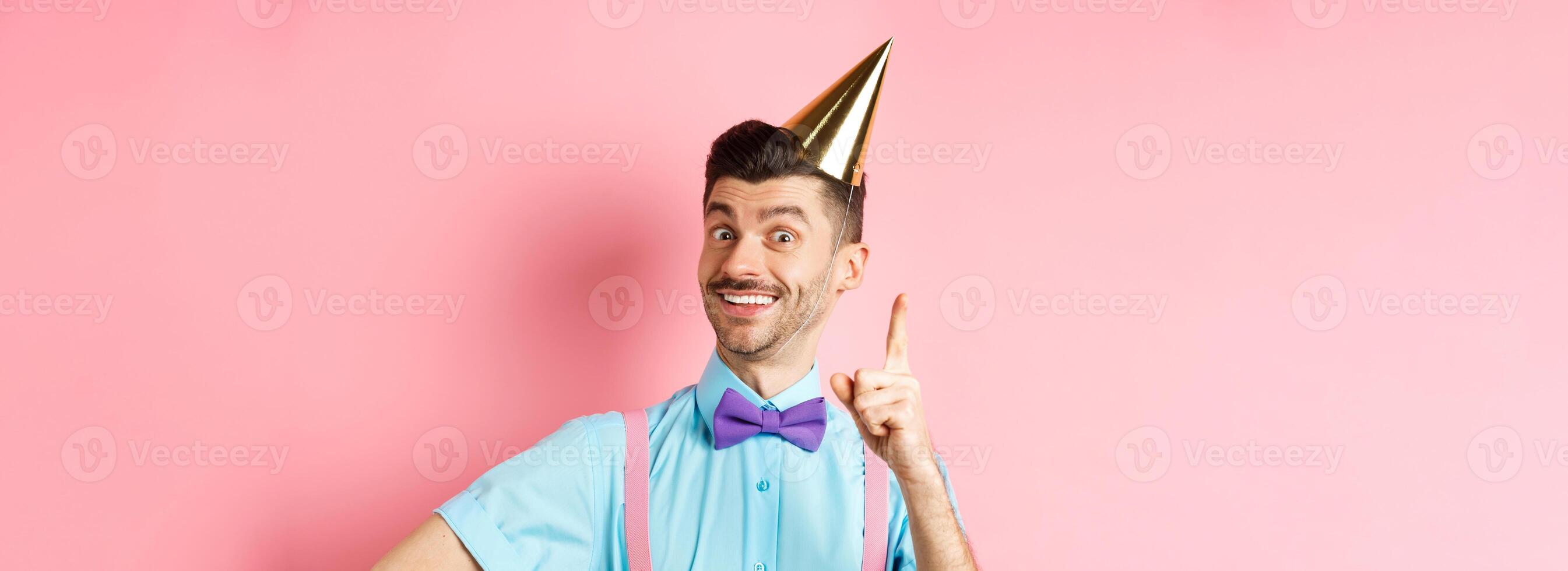 Holidays and celebration concept. Image of happy man in birthday party hat pitching an idea, raising finger and smiling, have plan or solution, standing over pink background photo