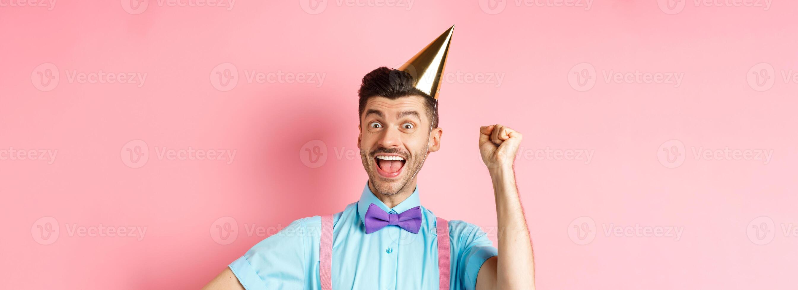 Holidays and celebration concept. Happy birthday boy with moustache and bow-tie celebrating event in party cone hat and festive clothes, raising hand up and shout yes photo