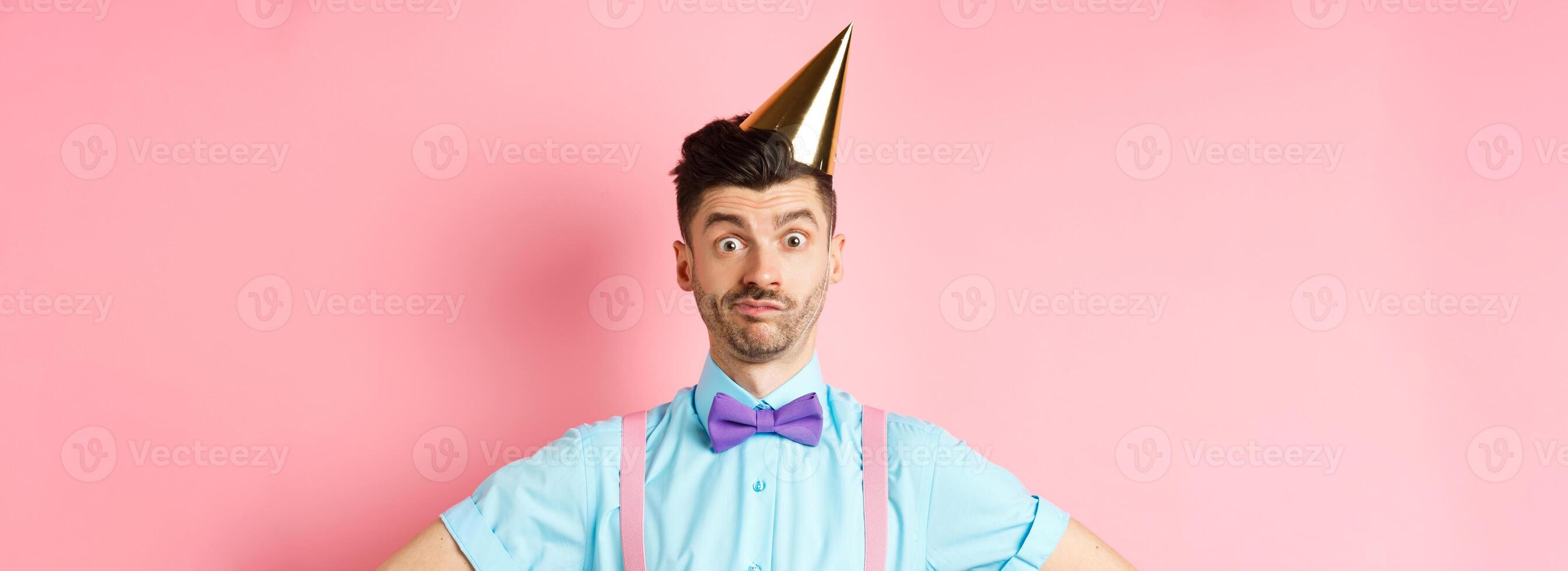 Holidays and celebration concept. Close-up of confused male entertainer in party hat and bow-tie, looking puzzled and shocked, standing over pink background photo