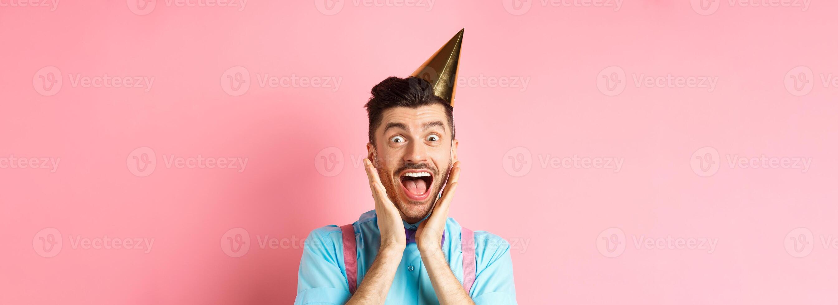 Holidays and celebration concept. Close-up of funny guy in birthday hat shouting surprised, screaming with joy, receiving awesome gift, standing happy on pink background photo