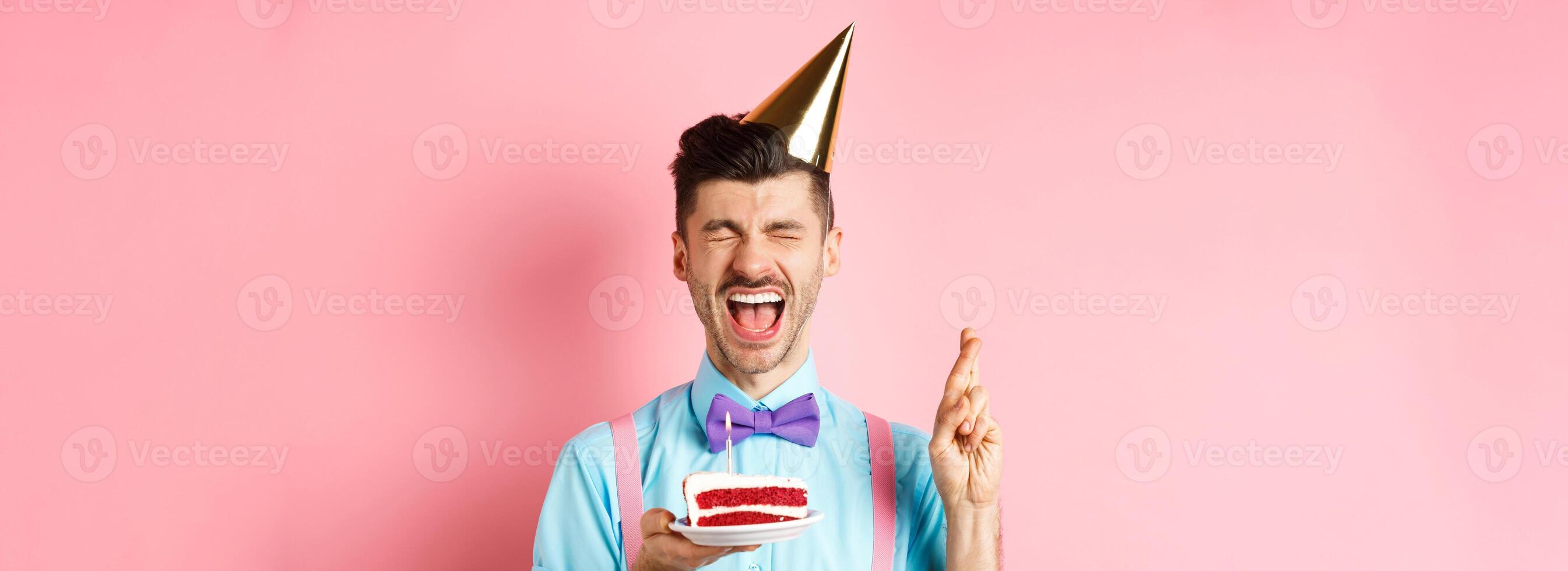Holidays and celebration concept. Excited guy celebrating birthday and making wish, cross fingers for good luck while holding bday cake with candle, pink background photo