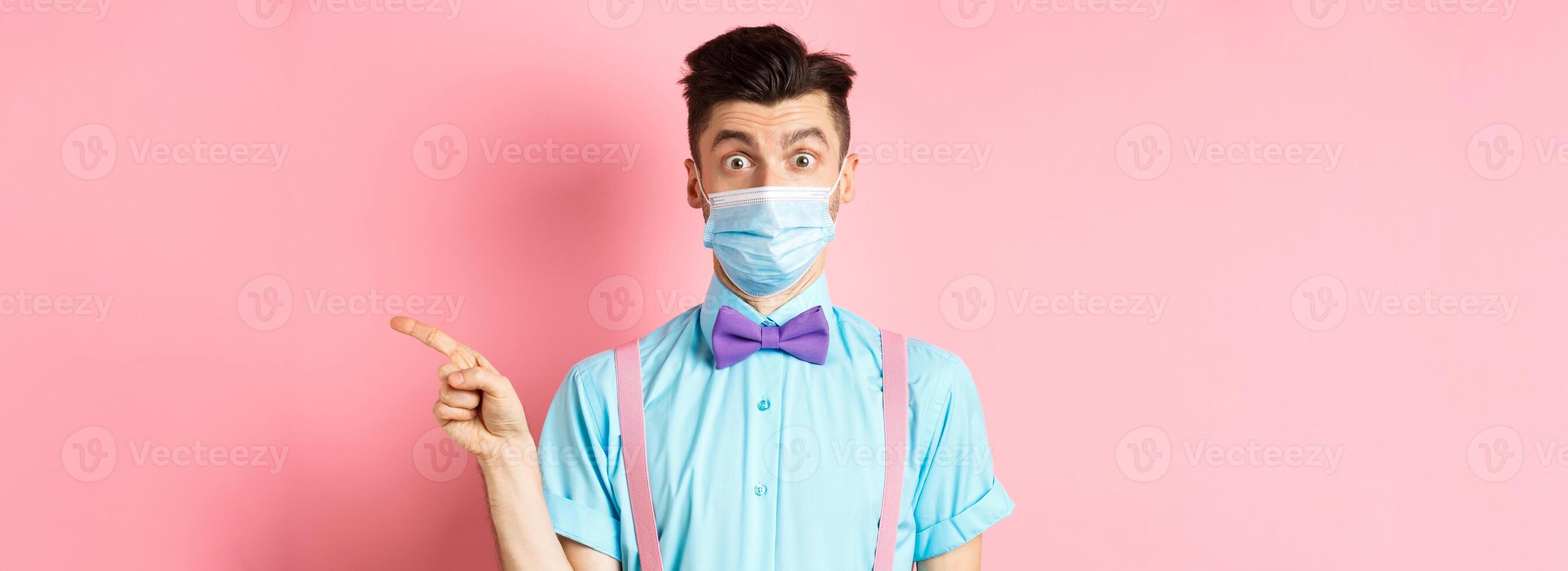 Coronavirus, healthcare and quarantine concept. Man looking surprised in medical mask, asking question and pointing right, curious about promo offer, standing on pink background photo