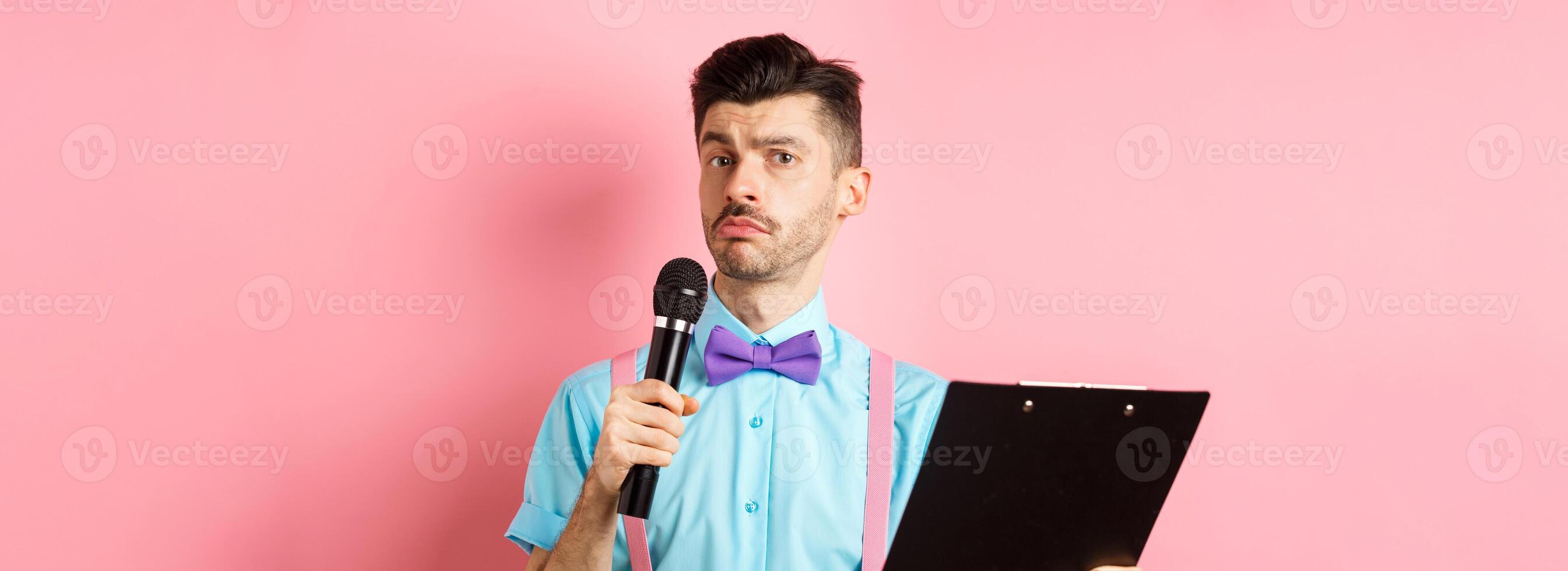 Holidays concept. Funny man with bow-tie making speech, perform on party events, holding script on clipboard and microphone, entertaining you, standing over pink background photo