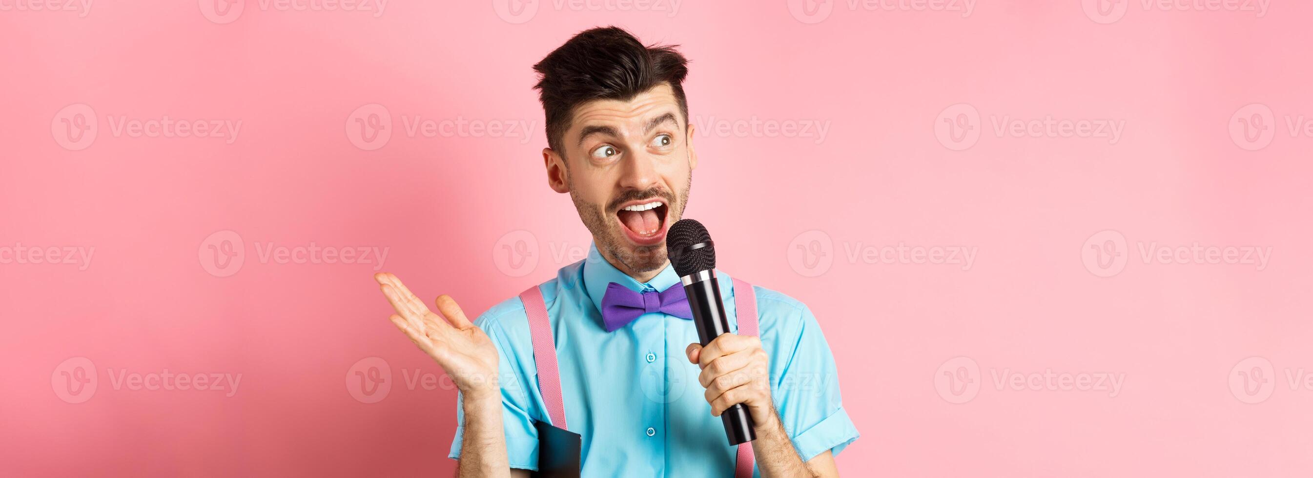 Image of male show host making speech, talking in microphone with clipboard under armpit, enteratin people on festive event, standing over pink background photo