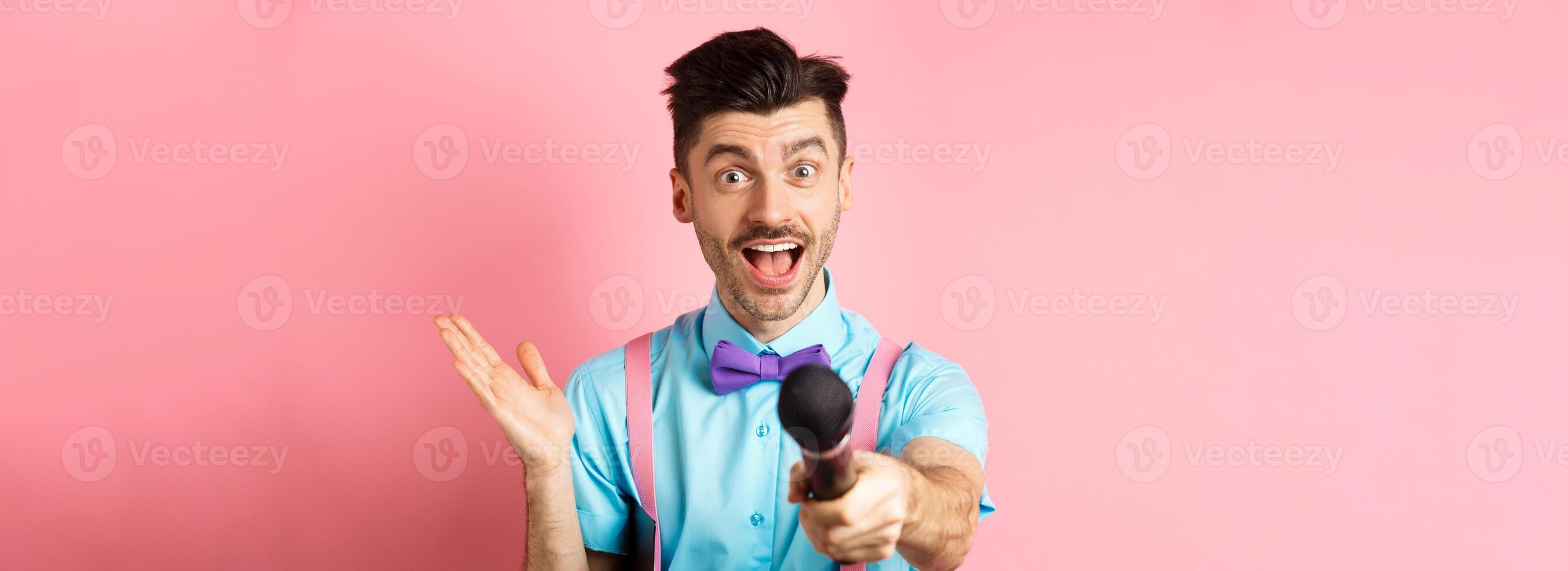 Cheerful young man, show host in bow-tie giving you microphone and smiling, entertain people on festive celebration events, performing at holidays, standing on pink background photo
