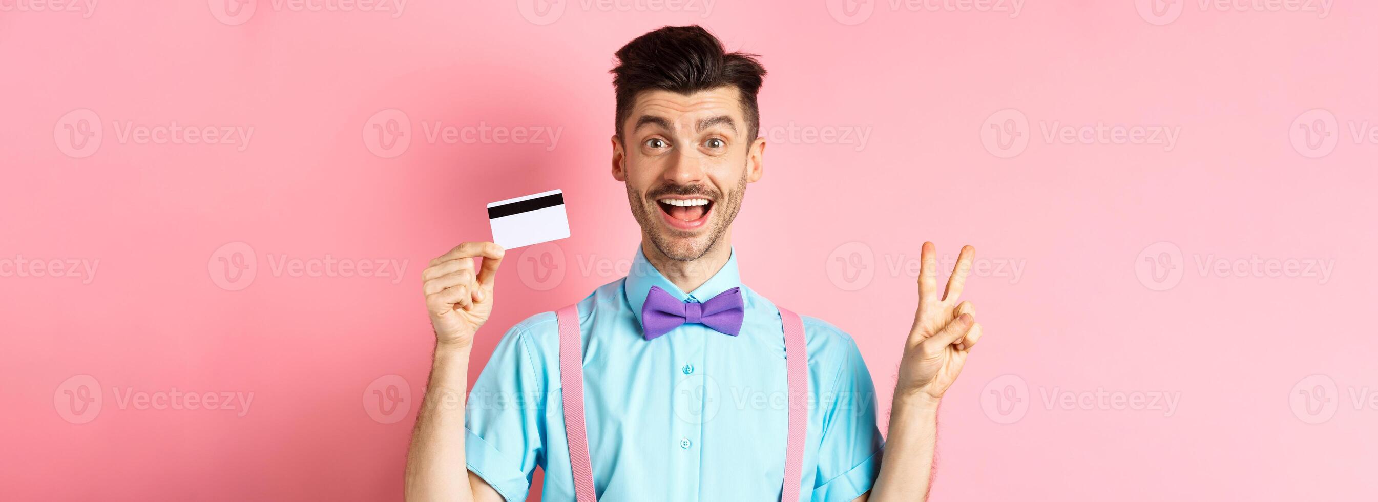 Shopping concept. Smiling caucasian guy in bow-tie showing plastic credit card and peace, victory sign, standing happy on pink background photo