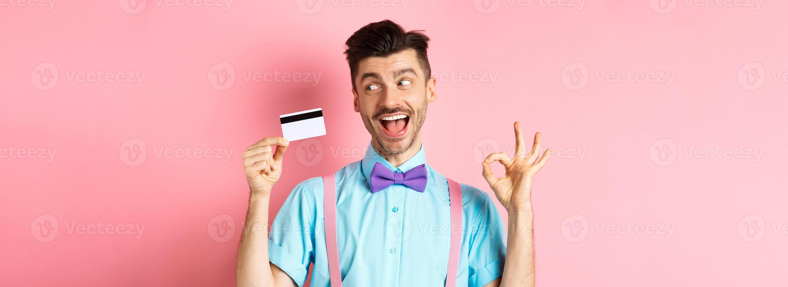 Cheerful man recommending great promo, showing OK sign and looking excited at plastic credit card, standing on pink background photo