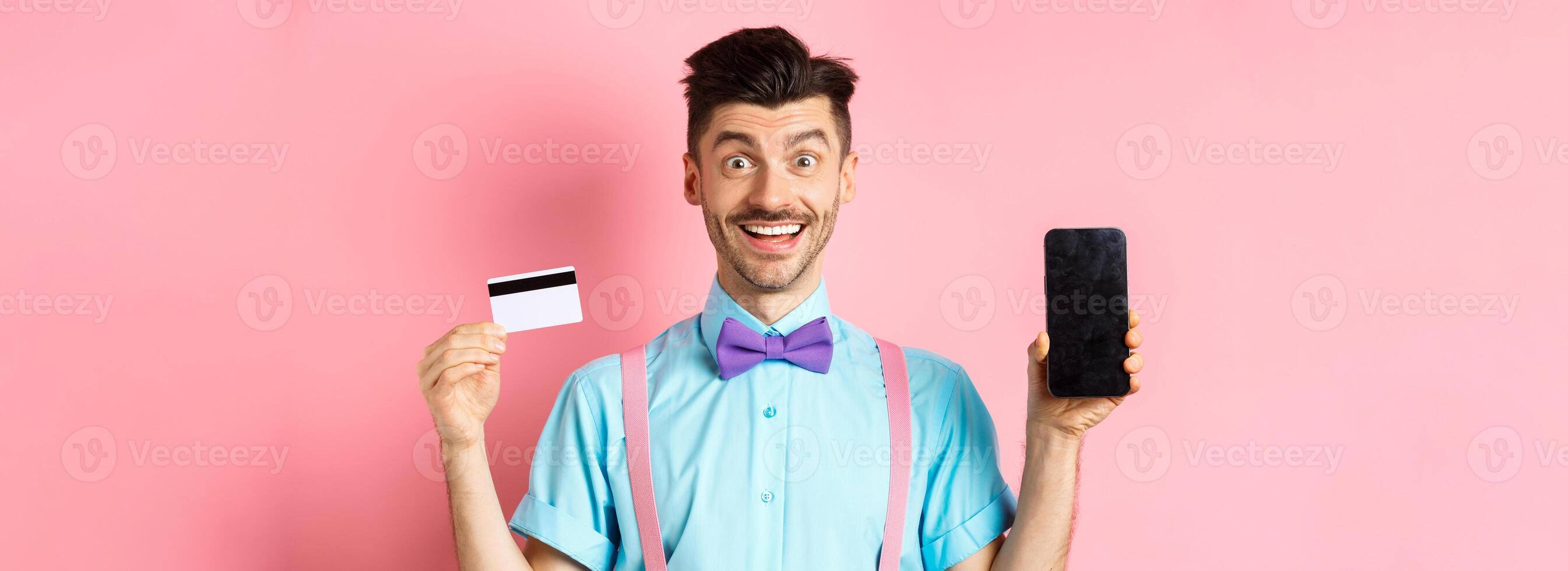 E-commerce and shopping concept. Happy man showing empty smartphone screen and plastic credit card, recommending app or online store, standing on pink background photo