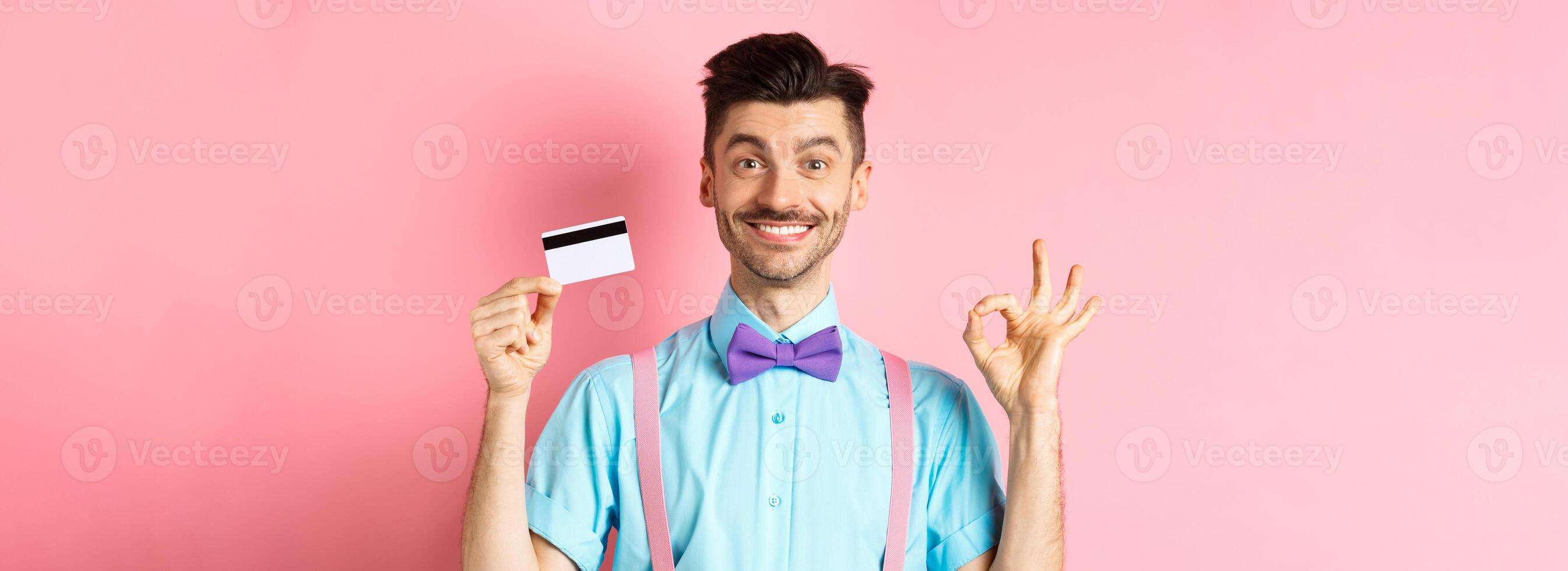 Shopping concept. Smiling handsome male shopper showing Ok sign and plastic credit card, buying something, standing satisfied on pink background photo