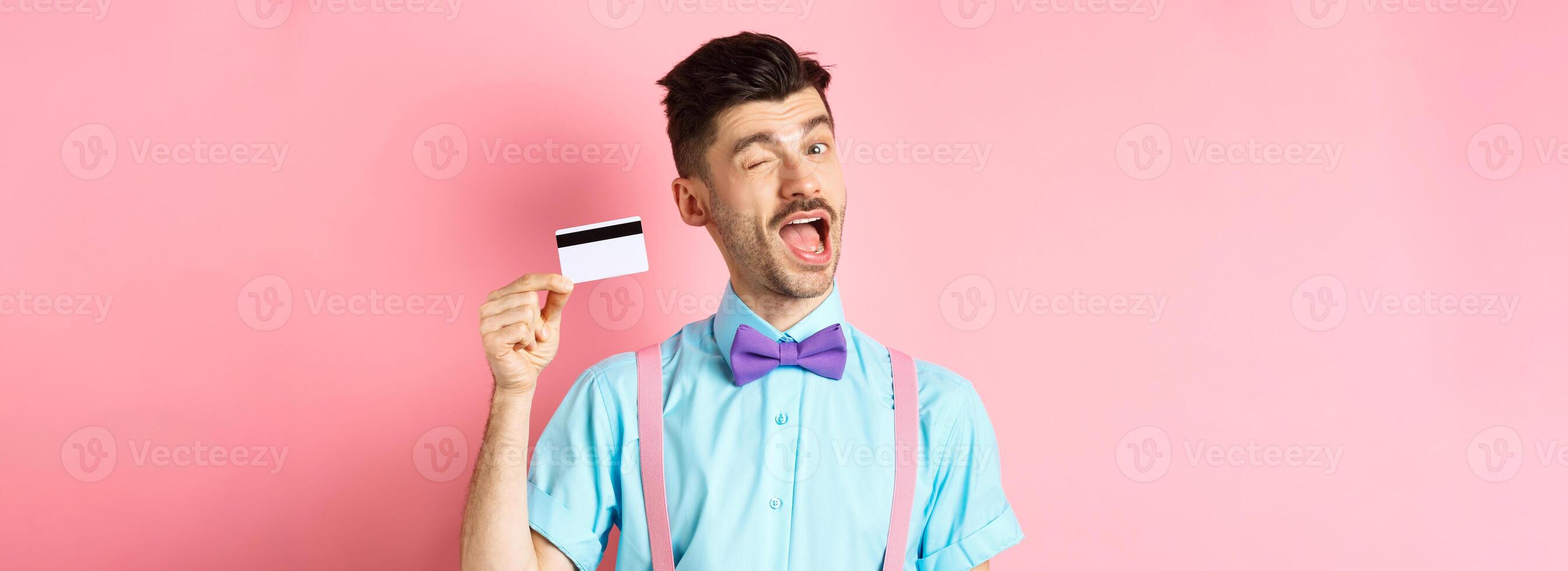 Shopping concept. Funny guy with moustache winking at camera, showing plastic credit card, recommending bank promo offer, standing on pink background photo