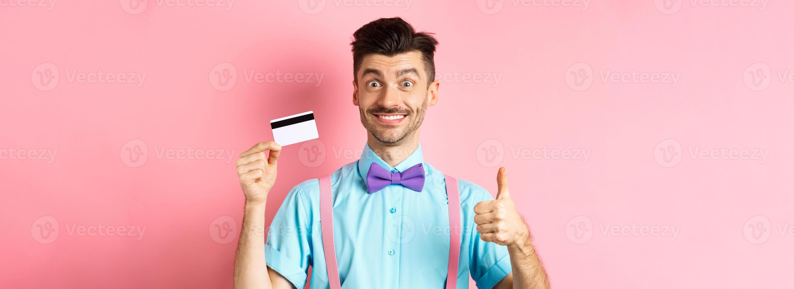 Cheerful guy in bow-tie showing thumb up and plastic credit card, like promo offer, smiling happy at camera, standing over pink background photo