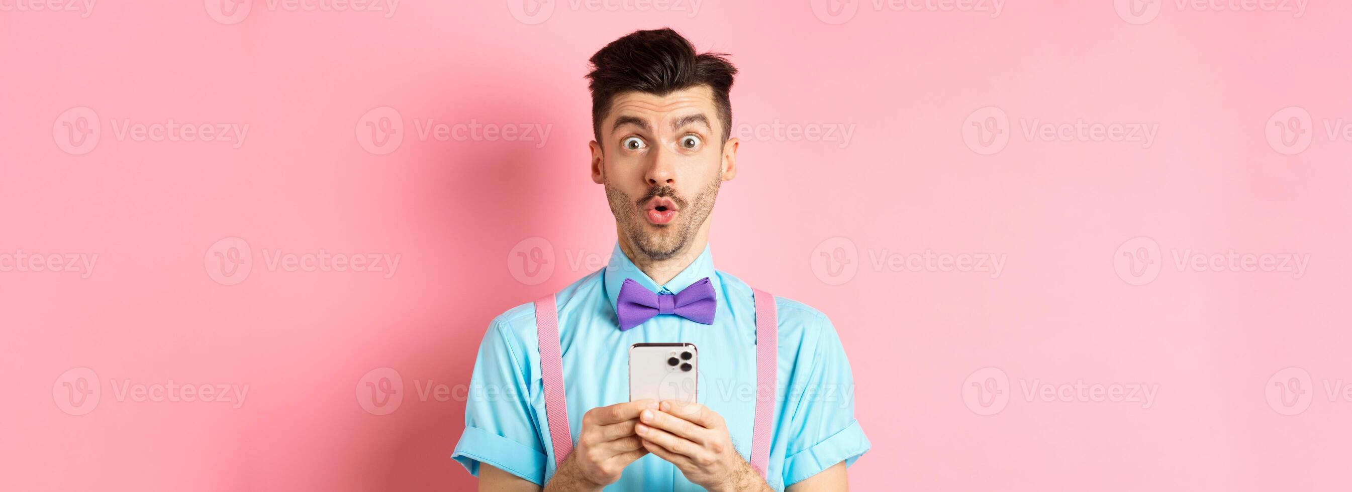 Online shopping. Intrigued guy in bow-tie, checking out internet promo offer on mobile phone, say wow at camera, holding smartphone, standing on pink background photo