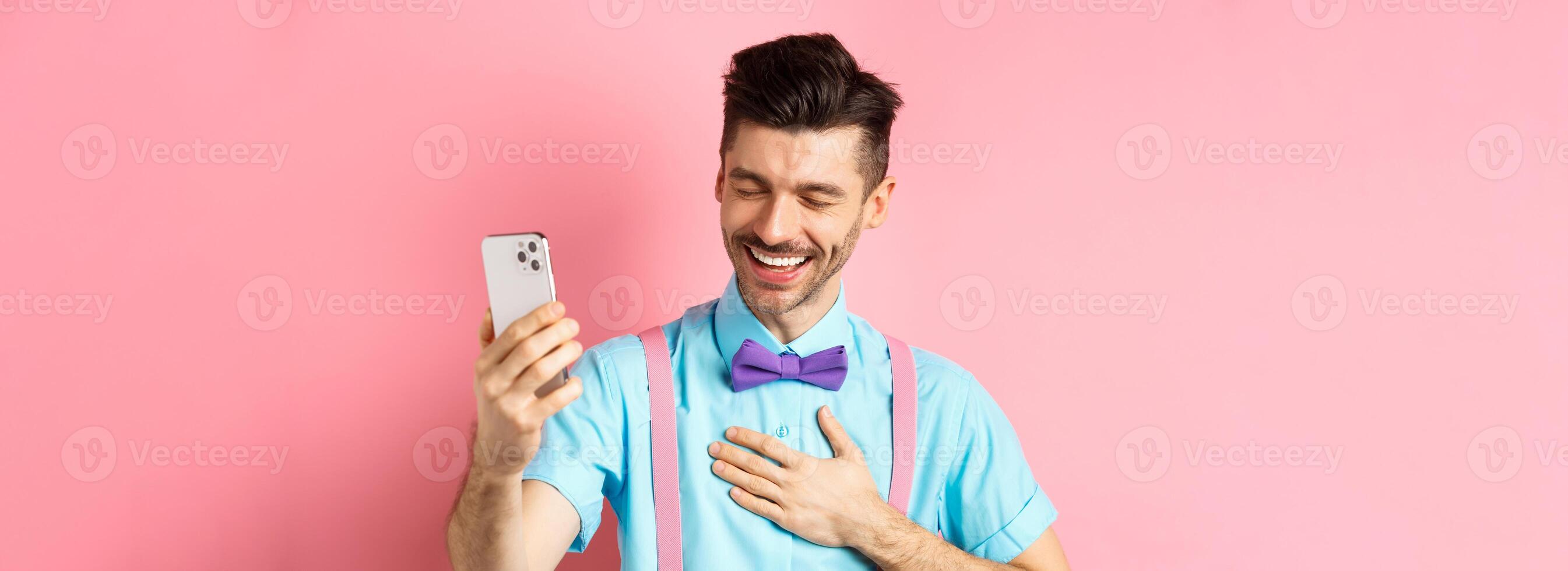Image of handsome guy laughing during chat, having mobile conversation and chuckle from something funny, standing on pink background photo