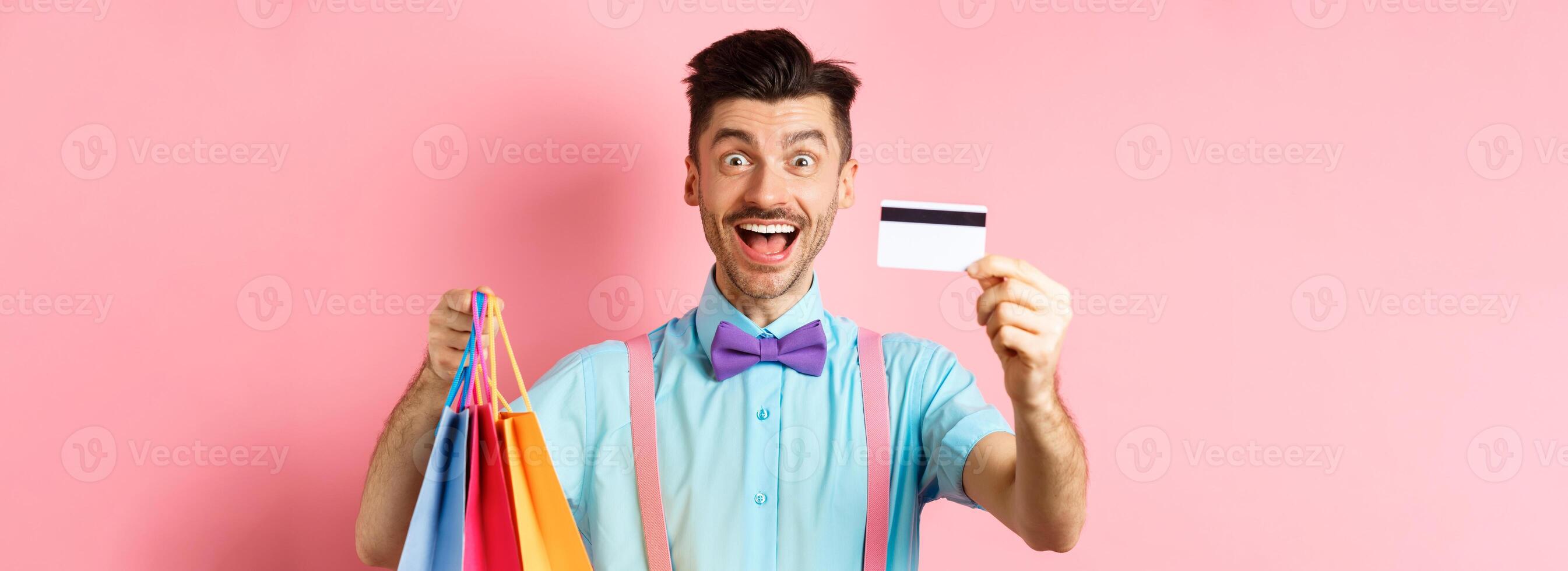 Happy young man giving his credit card and holding shopping bags, buying with discounts, standing on pink background and smiling photo
