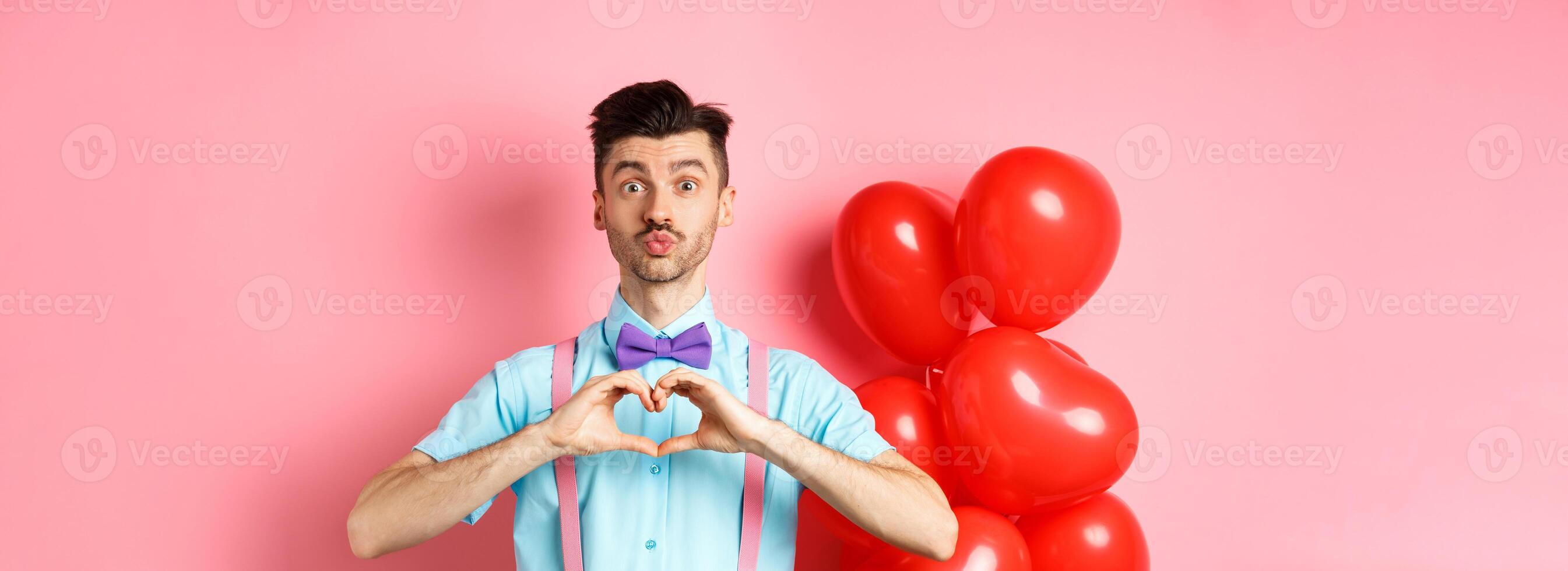 Valentines day concept. Cute boyfriend pucker lips for kiss, standing in bow-tie with heart gesture, say I love you, standing on pink background photo