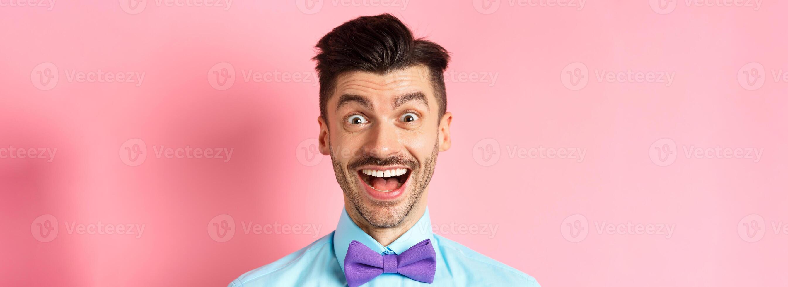 Close-up of surprised and happy young guy, open mouth and smiling amazed, checking out promo offer, standing in bow-tie over pink background photo