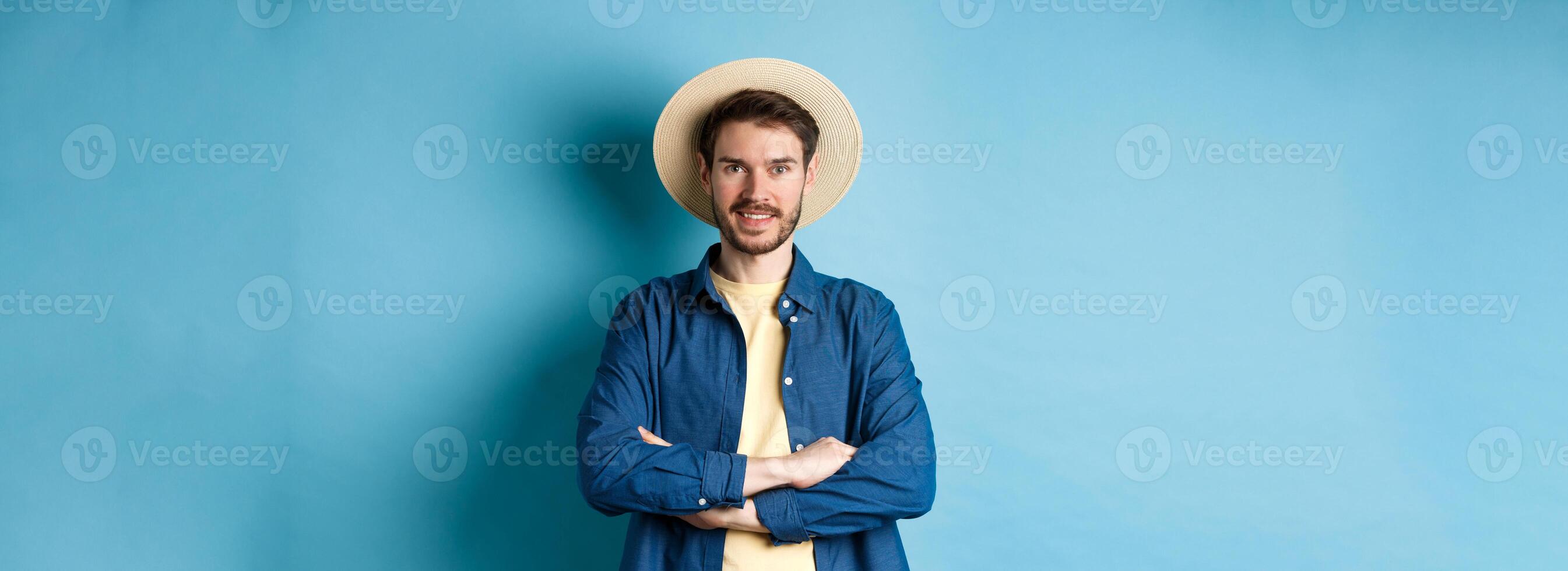 Portrait of cheerful guy going on vacation in straw summer hat, cross arms on chest and smiling, looking confident, standing on blue background photo