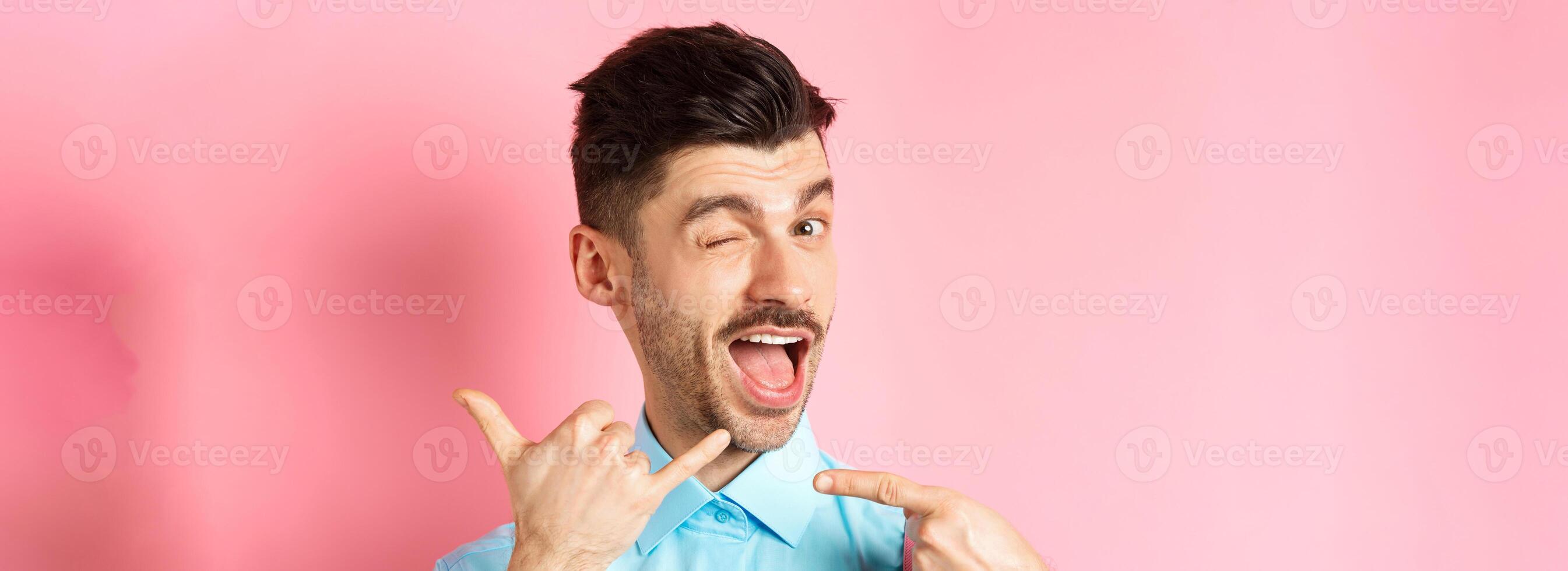 Cheeky and funny guy showing phone gesture, asking to call him and winking, telling dial his number, standing over pink background photo