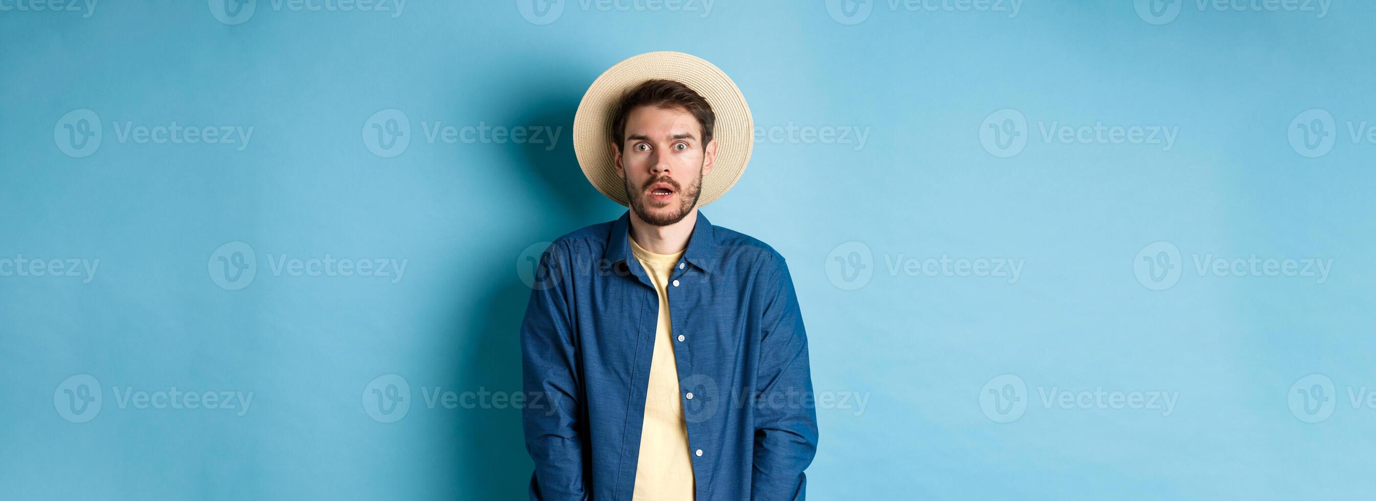 Young man tourist in summer hat drop jaw, staring at something shocking, standing on blue background photo
