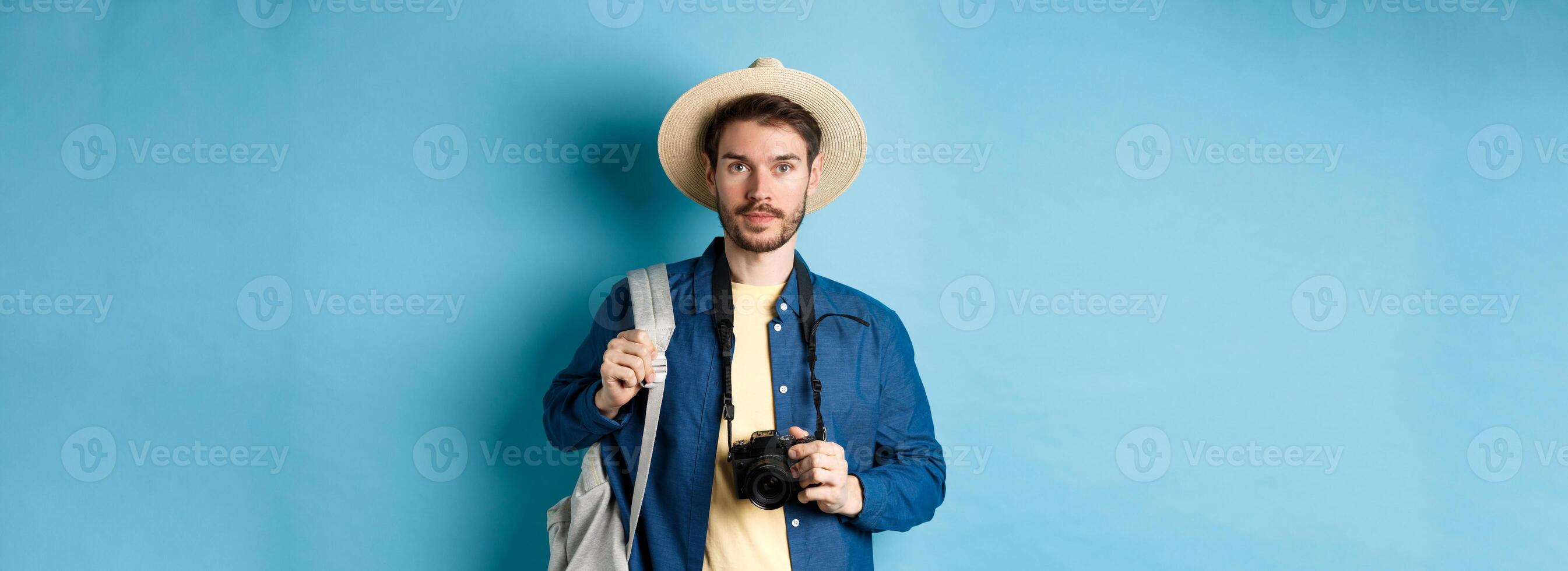 Handsome young tourist in summer hat, backpacking and travelling, holding photo camera, standing on blue background