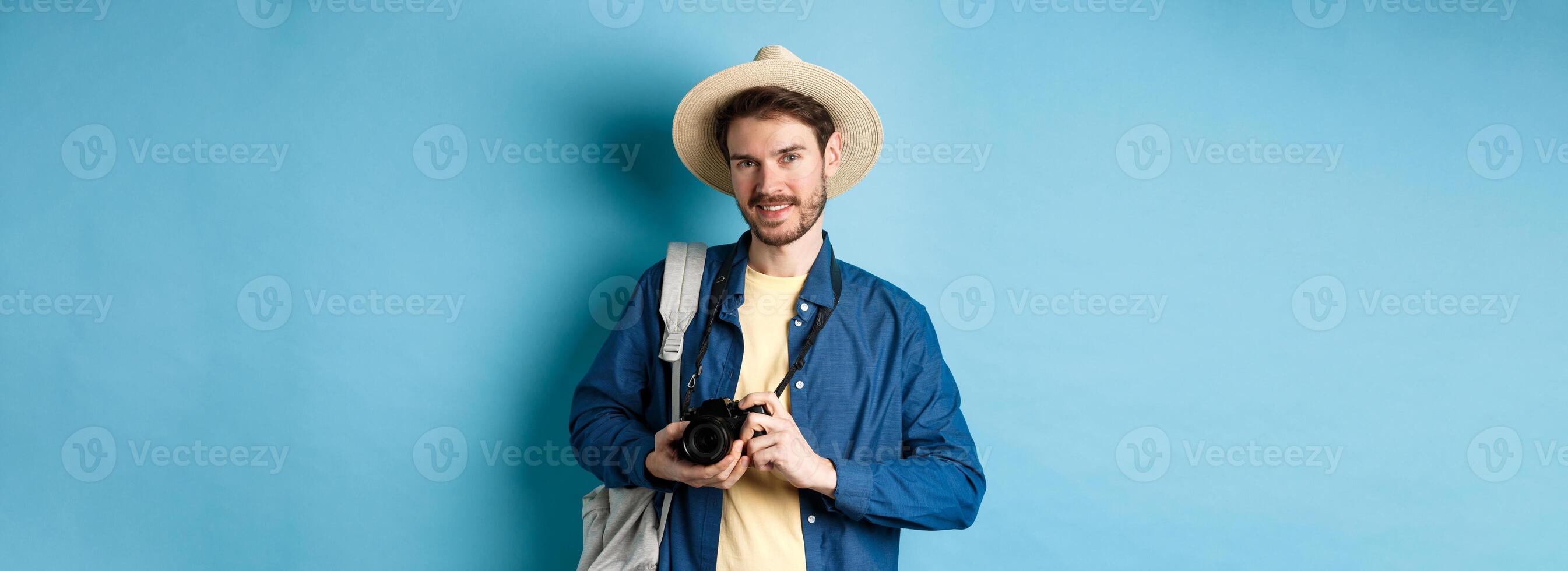 Handsome guy going on summer vacation, backpacking on holiday. Tourist with straw hat and camera smiling happy, standing on blue background photo