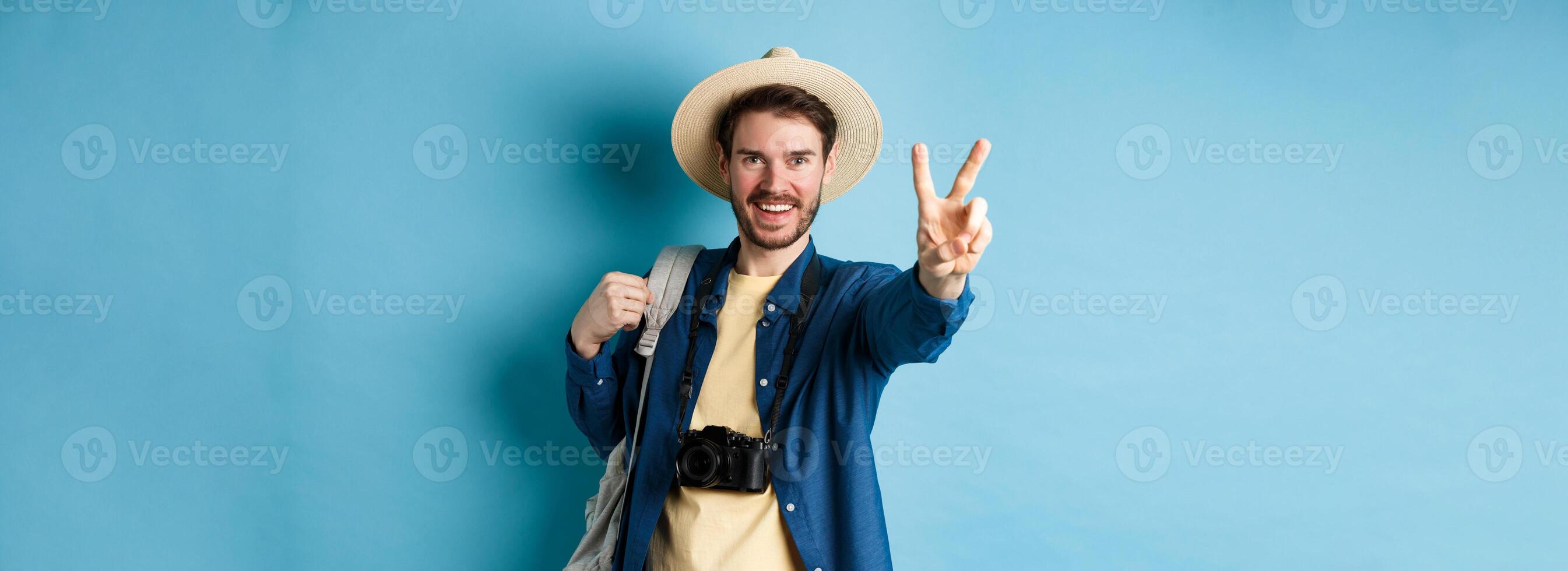 Handsome positive guy tourist showing peace sign on summer vacation, enjoying tropical holidays under sun, wearing straw hat, holding backpack and camera, blue background photo
