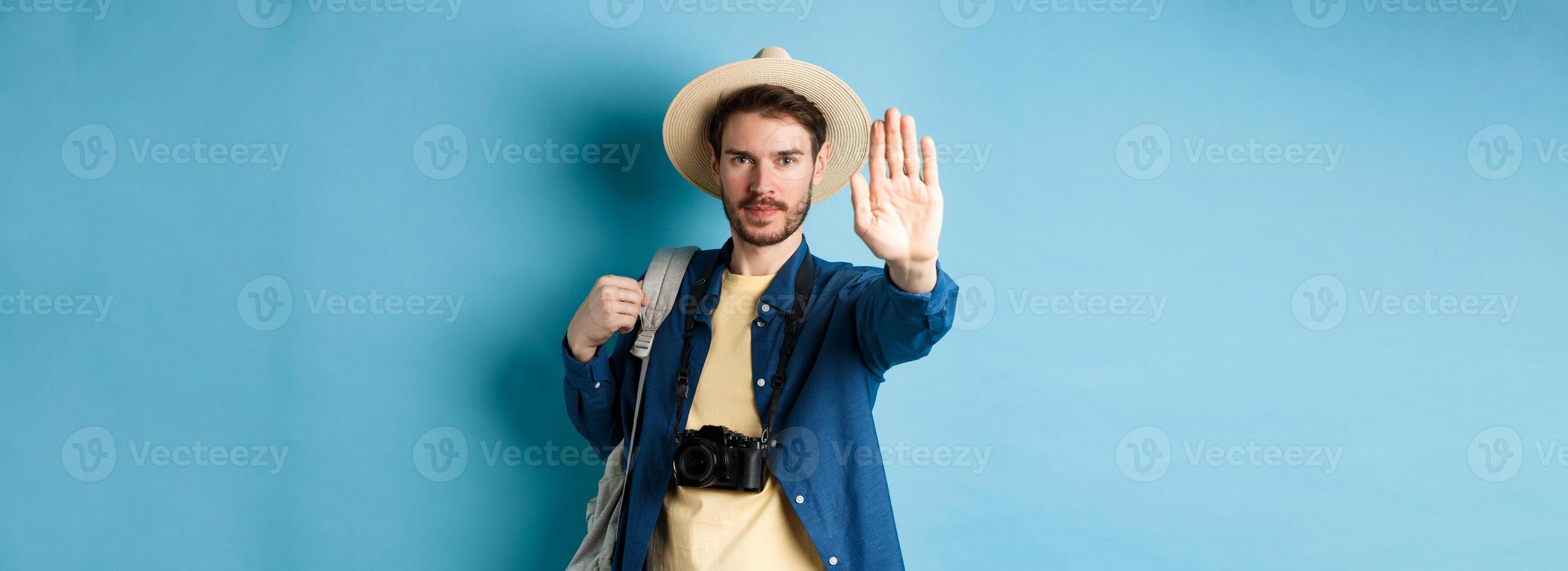 Serious male tourist warn you, stretch out hand to show stop gesture. Man on vacation forbid something, refusing or prohibiting action, standing with backpack on blue background photo