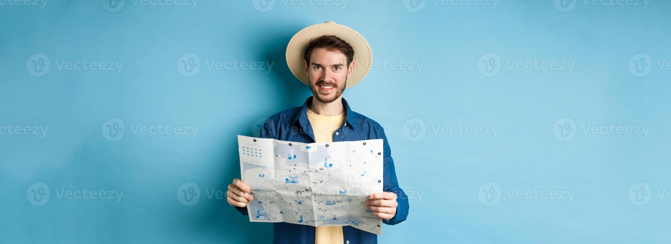 Happy and positive guy on vacation, looking at camera and holding map, smiling excited, going on summer travel, standing on blue background photo