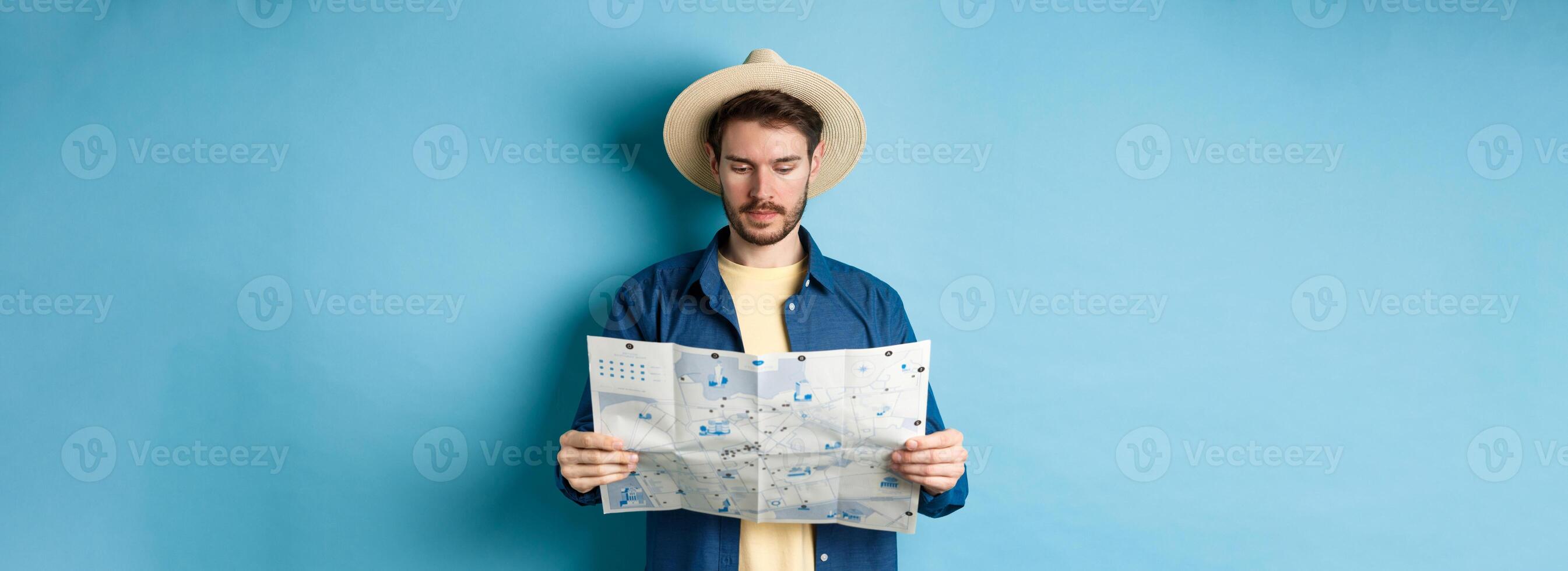 Handsome tourist with summer hat reading map, looking for sightseeing on vacation, standing on blue background photo