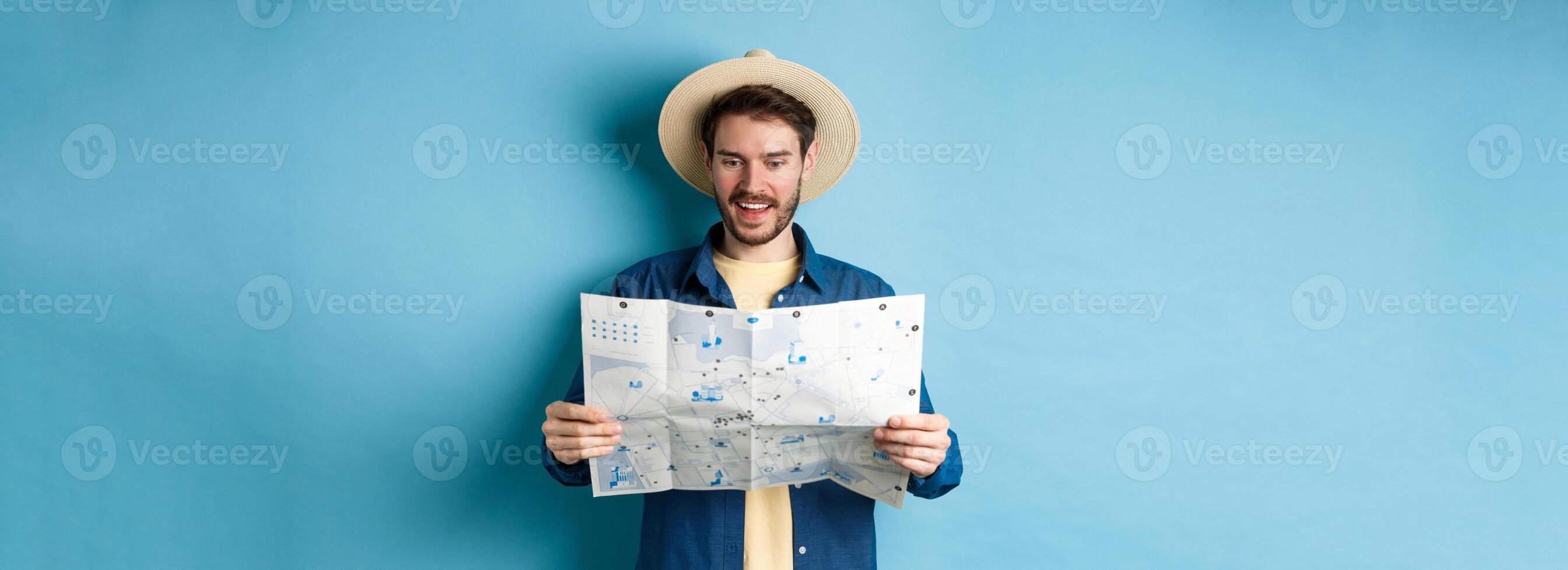 Cheerful tourist in summer hat planning travel route on vacation, looking at sighseeing map and smiling excited, standing on blue background photo