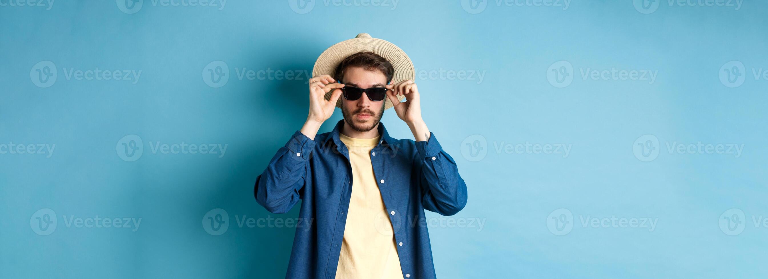 Handsome tourist in straw hat put on sunglasses on summer vacation, standing on blue background photo