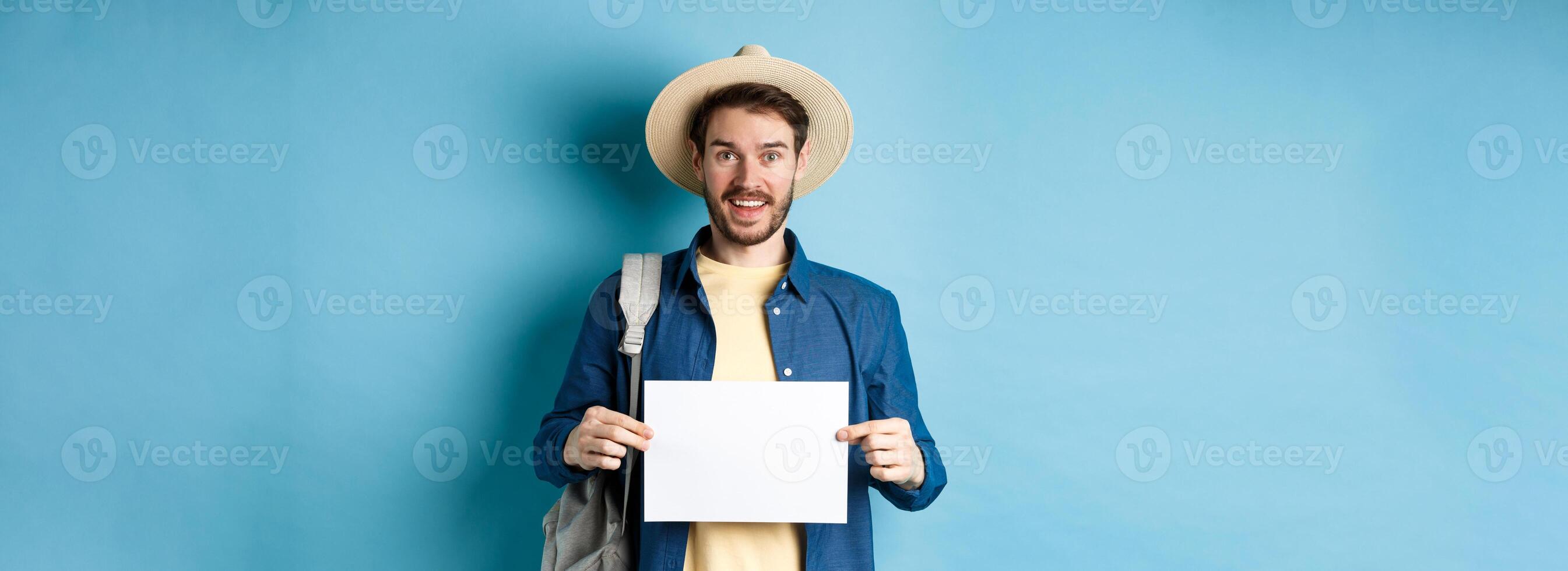 Cheerful tourist in summer hat, showing empty piece of paper and smiling, hitchhiking with backpack, standing on blue background photo