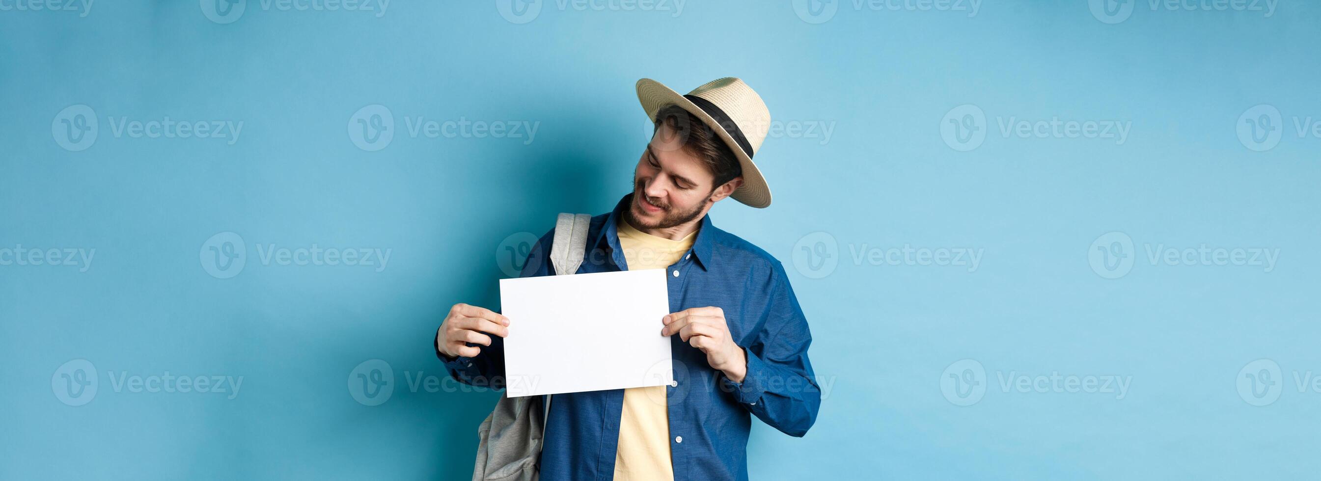 Happy tourist in straw hat smiling and looking at piece of paper for your logo, standing on blue background photo