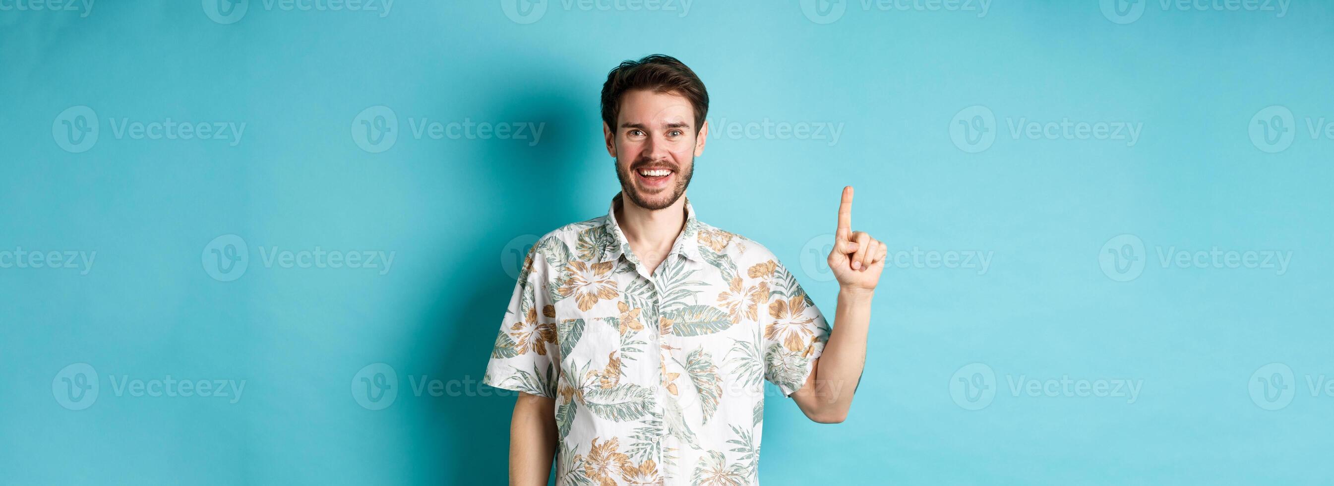 Happy tourist in hawaiian shirt smiling and pointing finger up, showing logo promotion, standing on blue background photo
