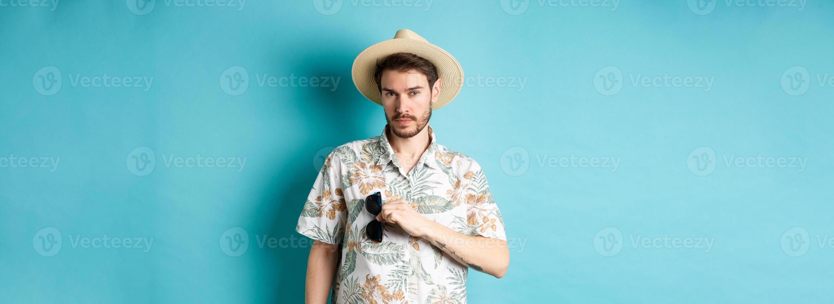 Handsome tourist in hawaiian shirt and summer hat, put sunglasses in pocket, going on vacation, standing against blue background photo