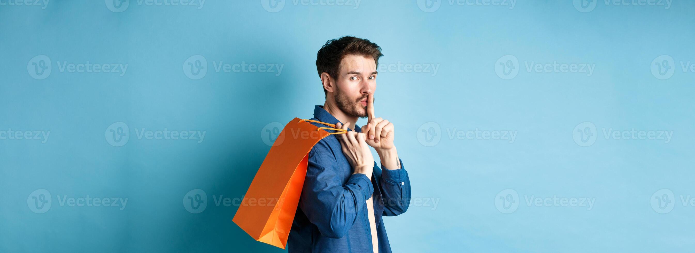 Image of modern guy holding shopping bag on shoulder, turn behind and shushing at camera, telling a secret, making surprise, standing on blue background photo
