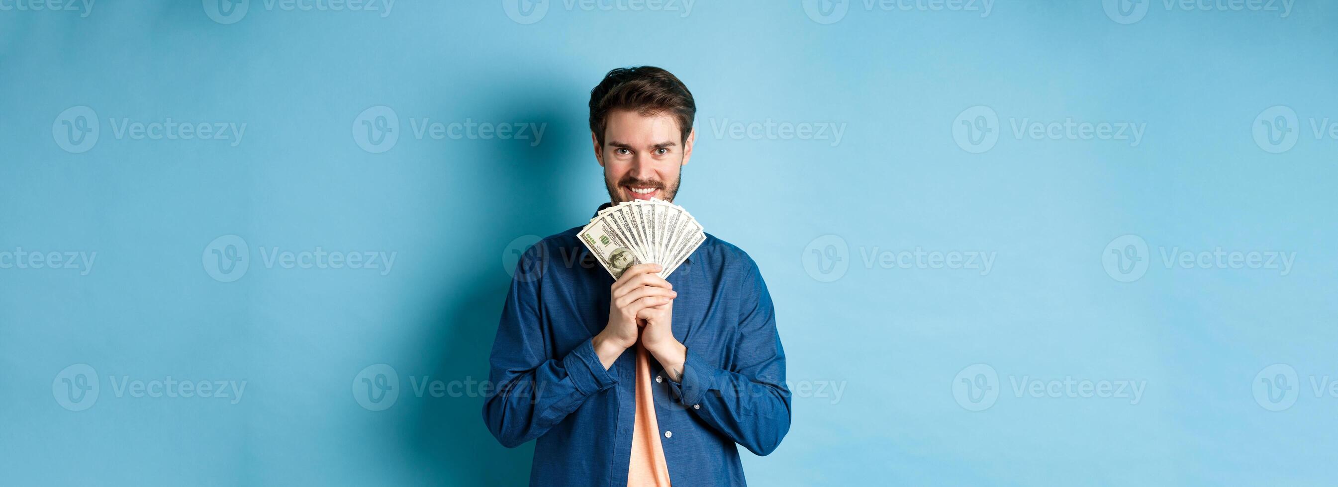 Happy smiling man holding money and looking at camera pensive, thinking of shopping, standing on blue background photo