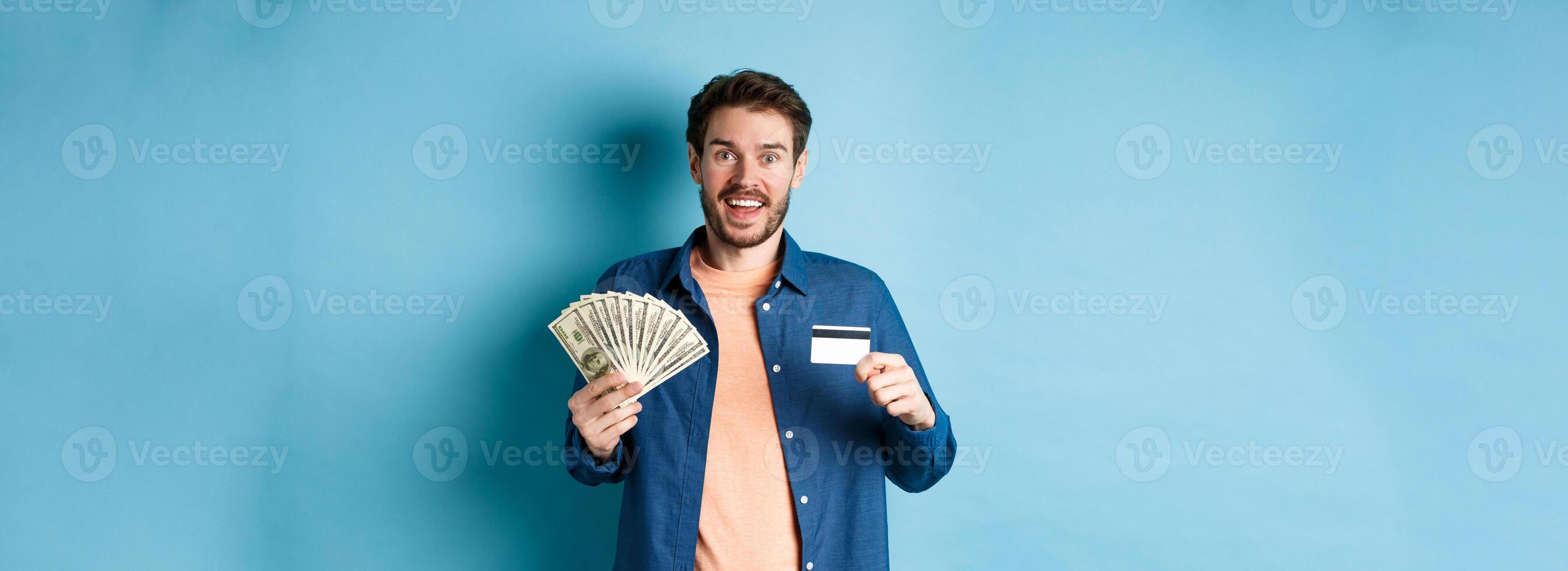 Smiling caucasian guy showing plastic credit card and money, looking happy, standing on blue background photo