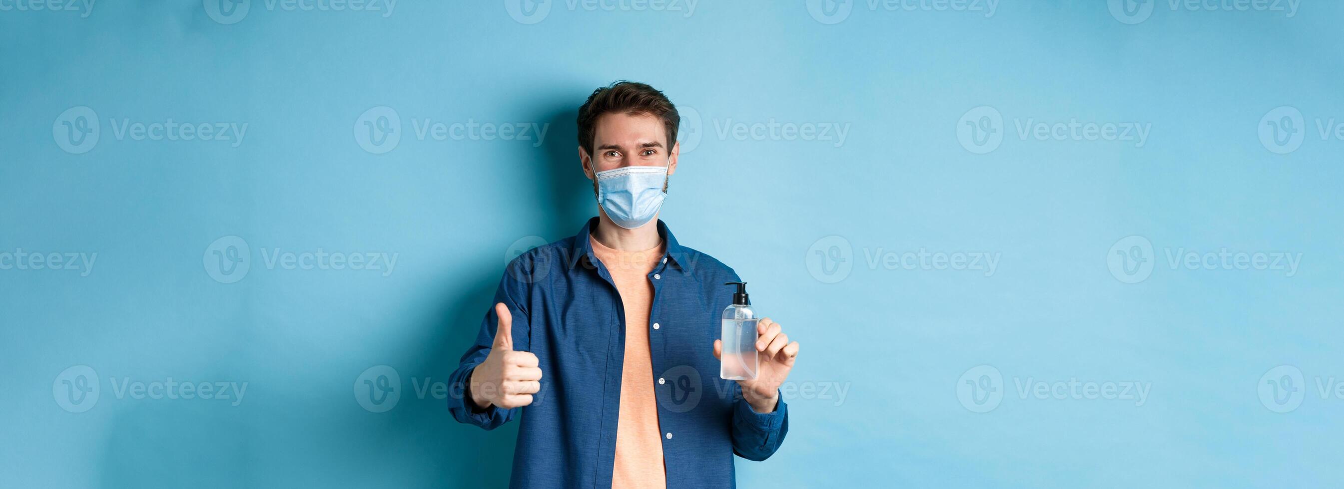 Coronavirus, quarantine and social distancing concept. Happy male model in face mask showing hand sanitizer and thumb up, praise good product, blue background photo
