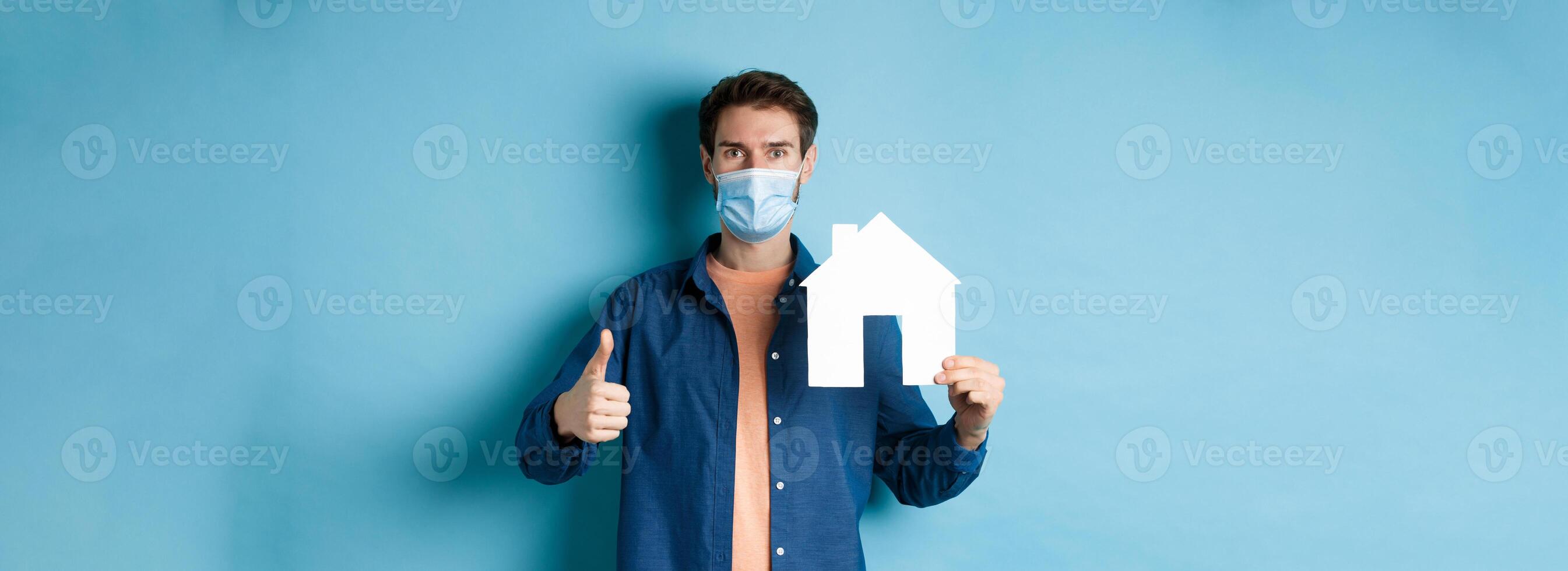 Real estate and covid concept. Young man in face mask showing paper house cutout and thumb-up, recommending agency, standing on blue background photo
