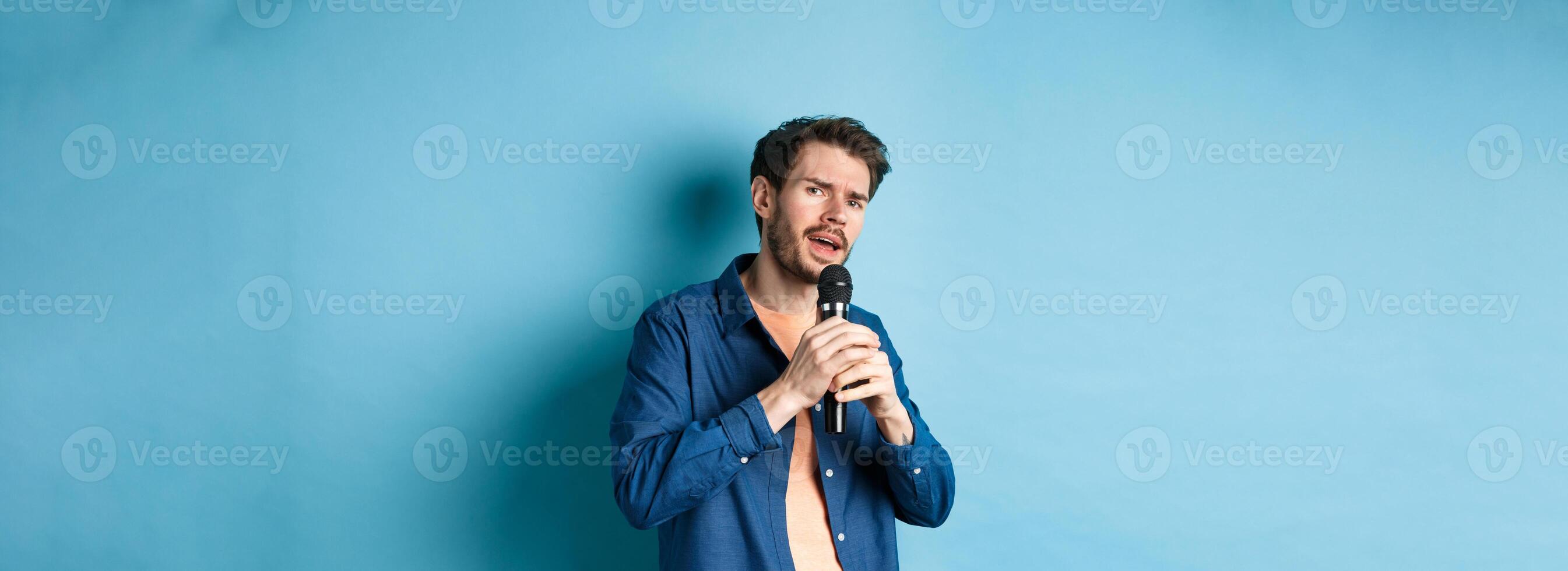 Young man singing song in microphone, playing karaoke, standing on blue background photo