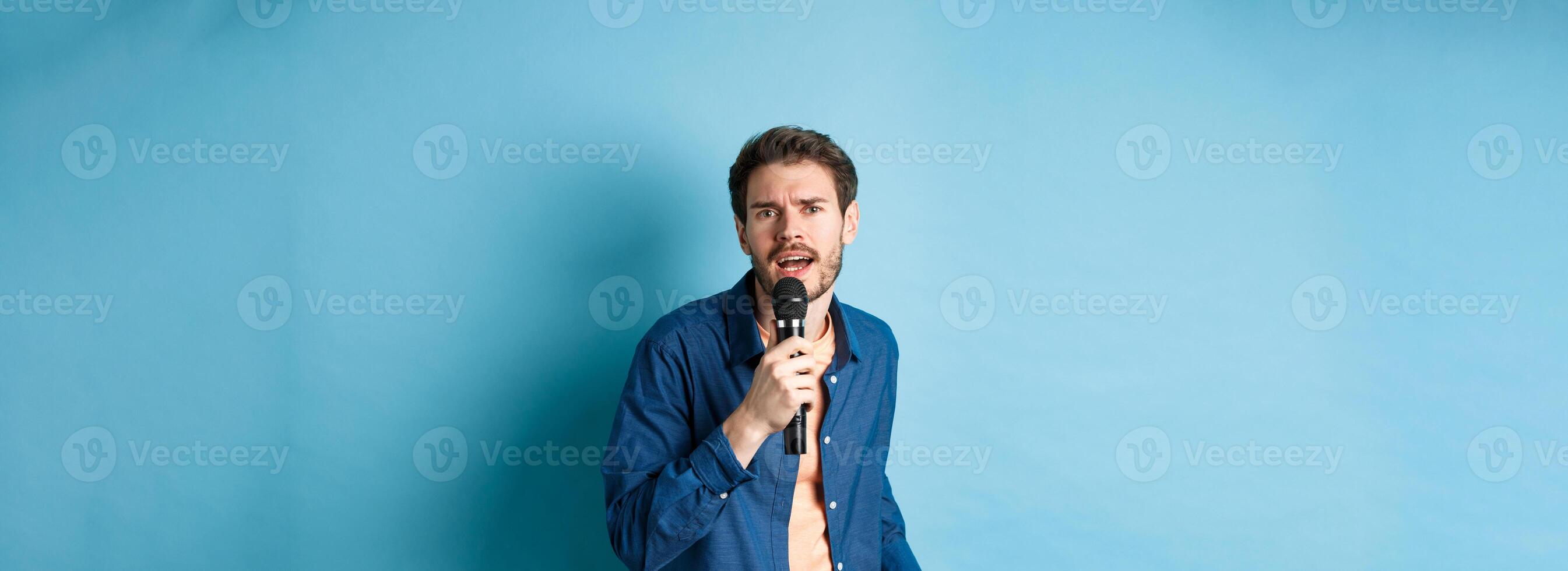 Passionate singer looking at camera, singing in microphone, playing karaoke on blue background photo