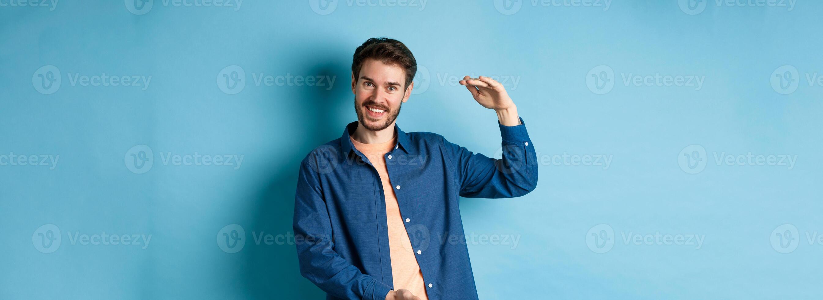 Handsome caucasian guy showing something big, shaping large size object with hands and smiling, standing on blue background photo