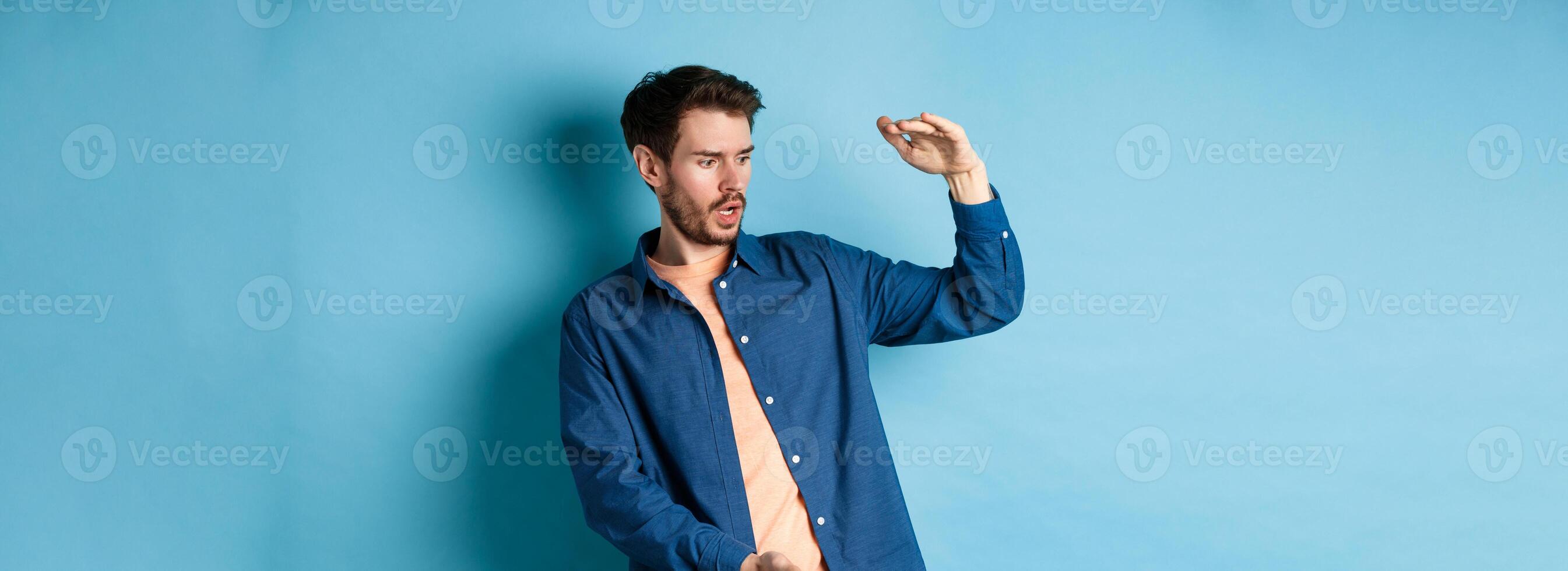 Shocked young man looking at empty space and showing big size object, standing on blue background photo
