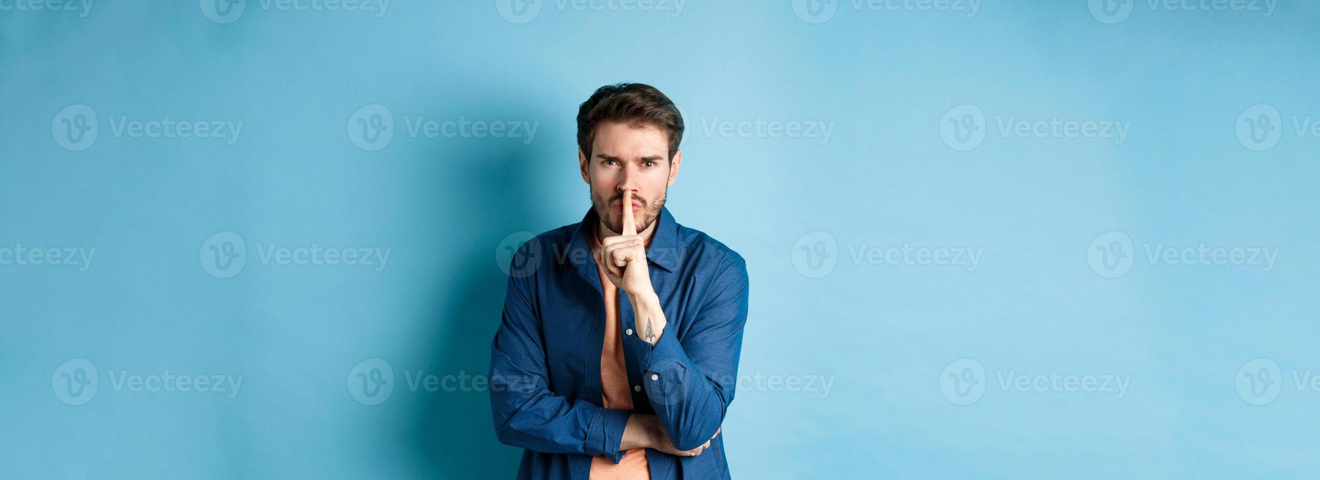 Angry man shushing, press finger to lips and frowning, tell to be quiet, show taboo gesture, standing on blue background photo