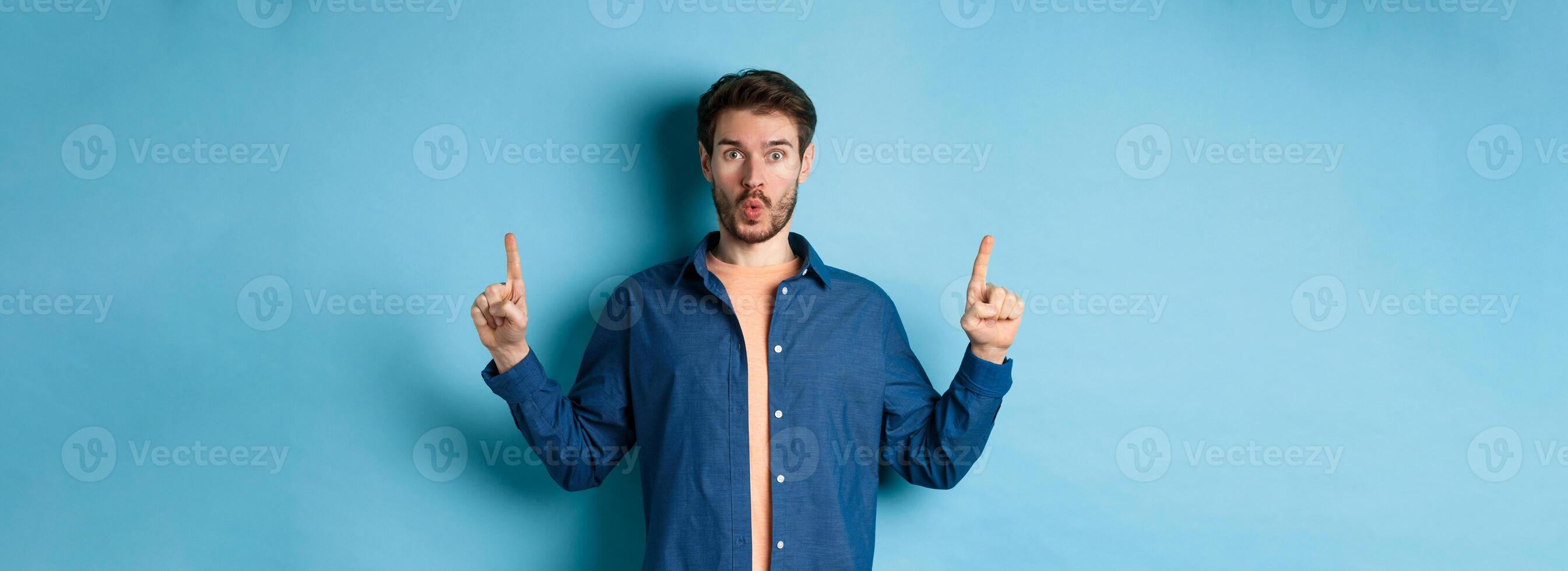 Surprised young man say wow, checking out advertisement, pointing fingers up and looking impressed, standing on blue background photo